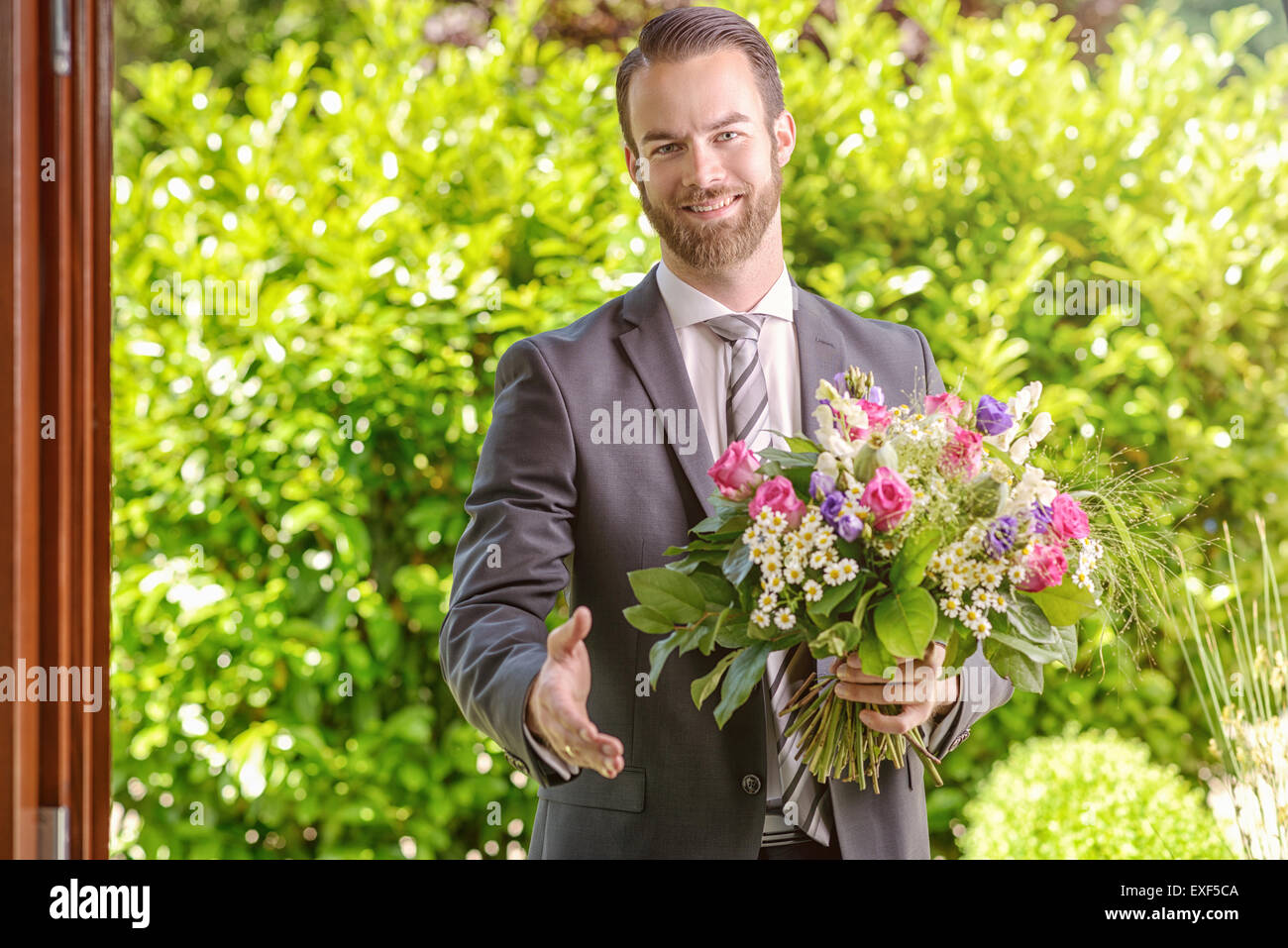 Handsome Young Businessman Holding a Bouquet of Fresh Flowers, Showing Handshake Gesture While Smiling at the Camera. Stock Photo