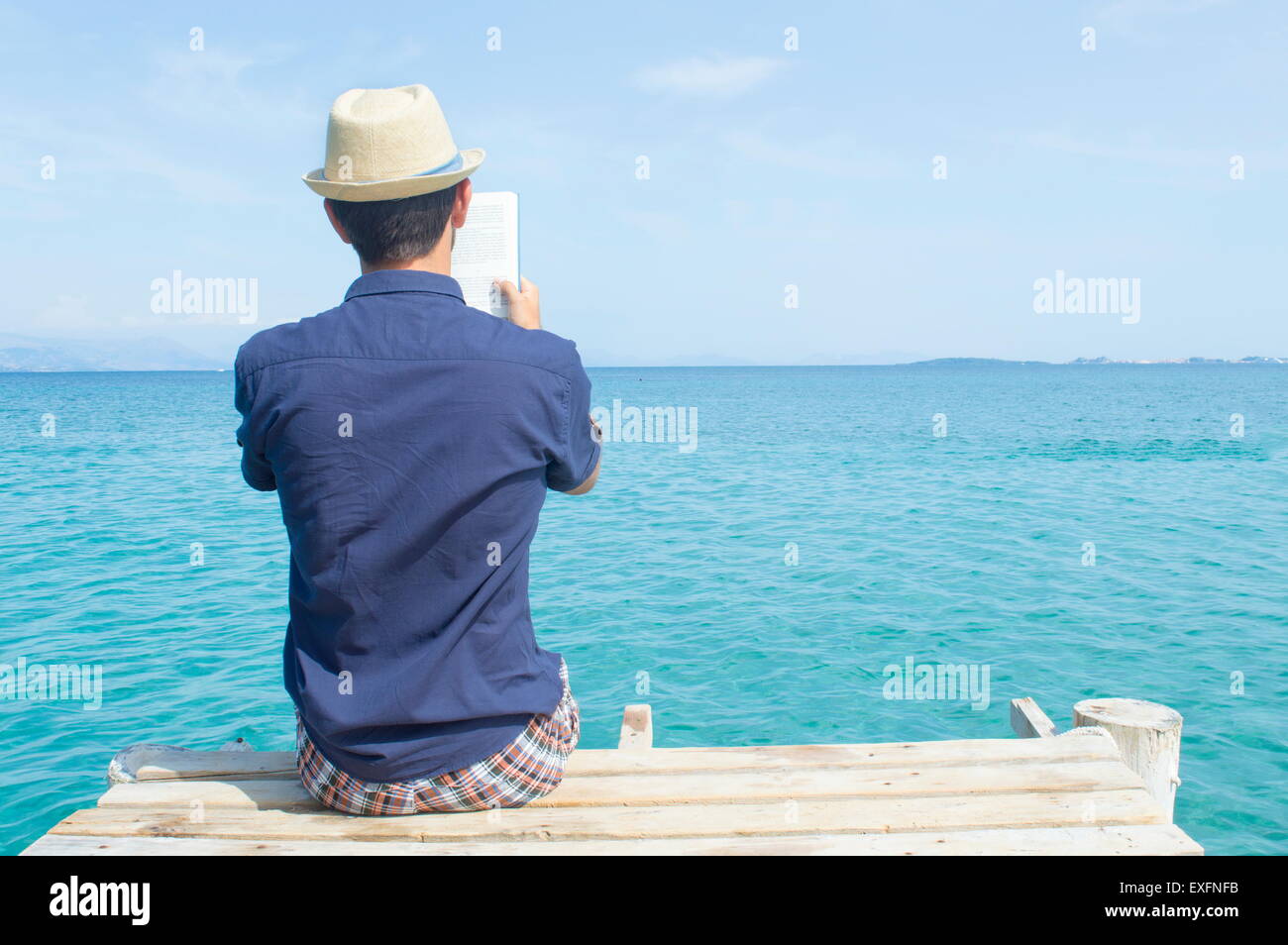 Young man in blue shirt sitting on the dock reading a book Stock Photo