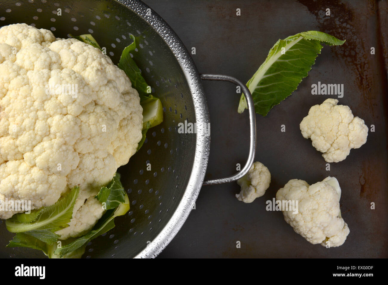 High angle closeup of a colander with a head of cauliflower. On the surface outside are a couple florets and a leaf. Stock Photo
