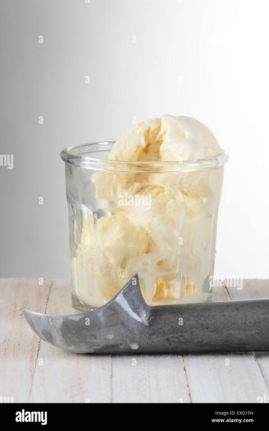 Glass of Vanilla Ice Cream and scoop on a wood table with a light to dark gray background. Stock Photo