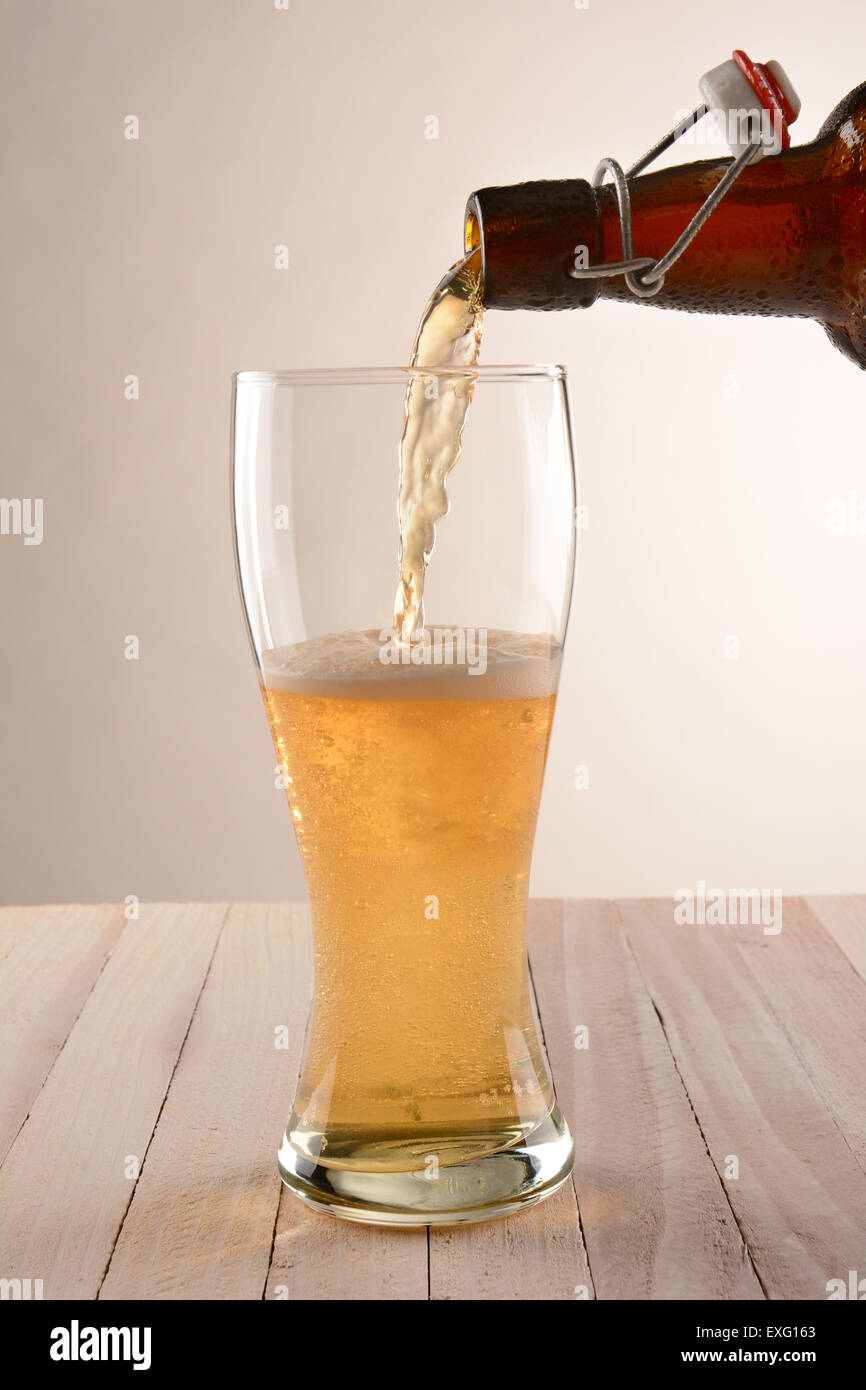 A brown swing top bottle of beer pouring into a glass. The partially filled glass is on a wood table with a light to dark Stock Photo