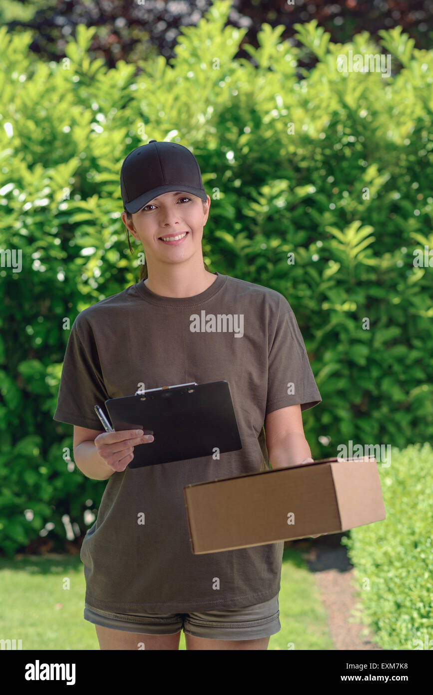 Smiling attractive female courier in a cap delivering a parcel to the door of a house holding out a clipboard for signature to a Stock Photo