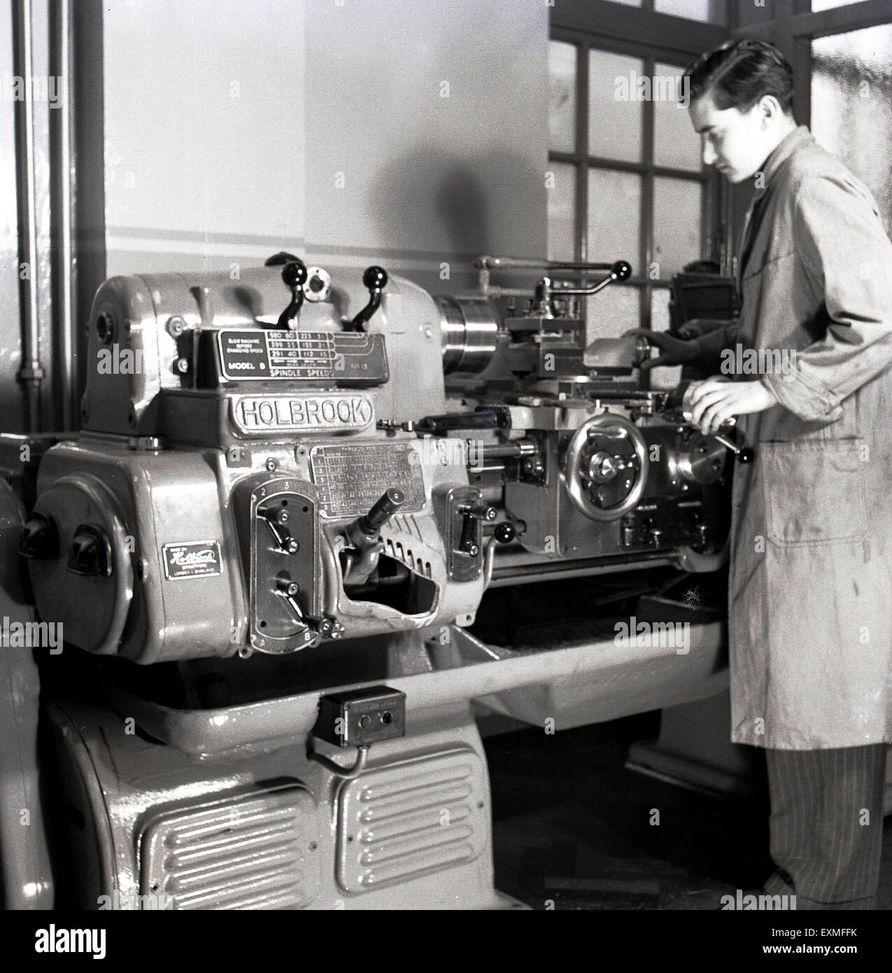 1950s, historical picture showing a young adult male using a sophiscated precision engineering machine tool, a mechanical bench lathe made by the Holbrook Machine Tool Co, a British company founded in 1850. In 1917 it started making self-contained motor-driven lathes and later manufactured precision optical chasing lathes, of which a large version is seen in the picture. Stock Photo