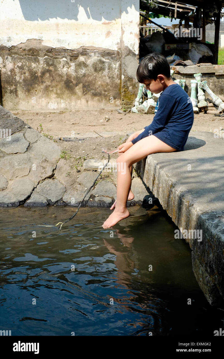 little boy sitting alone on small stone bridge over waterway playing fishing with a piece of string in a village near solo java Stock Photo