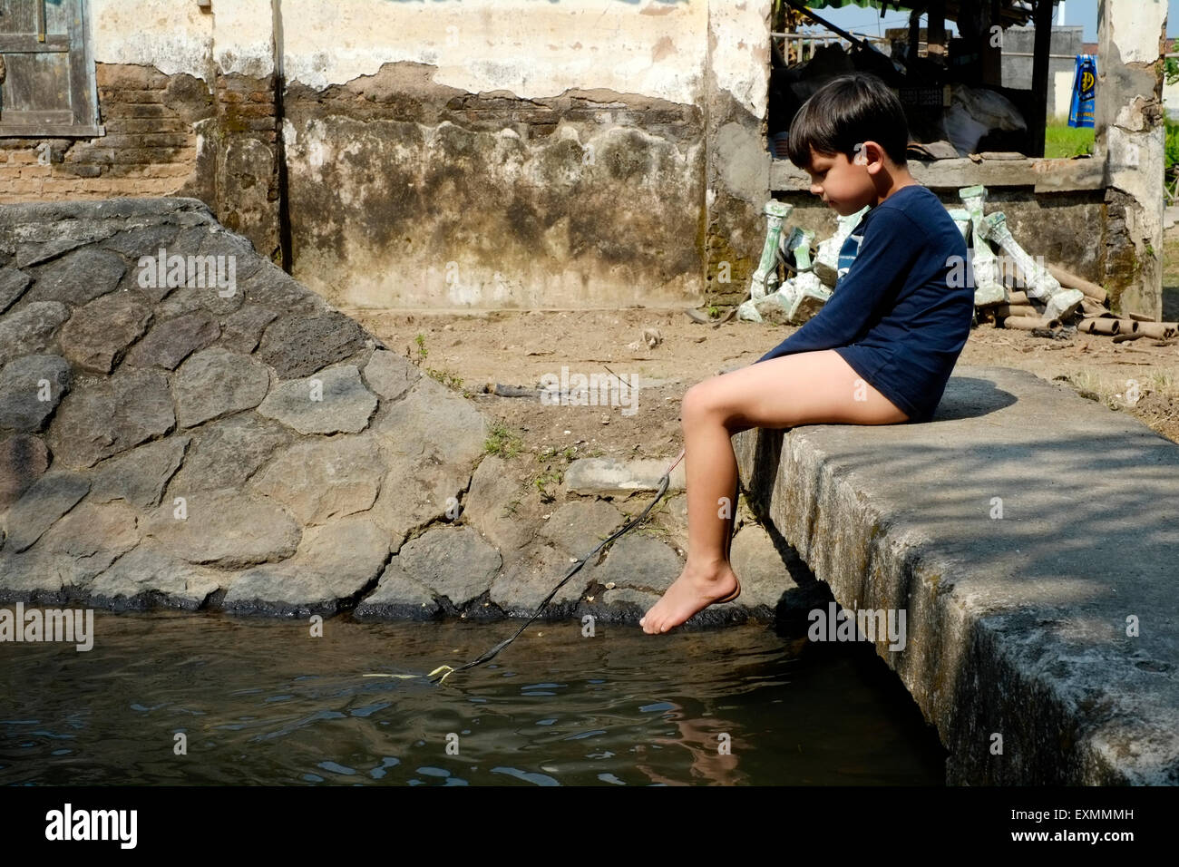 little boy sitting alone on small stone bridge over waterway playing fishing with a piece of string in a village near solo java Stock Photo