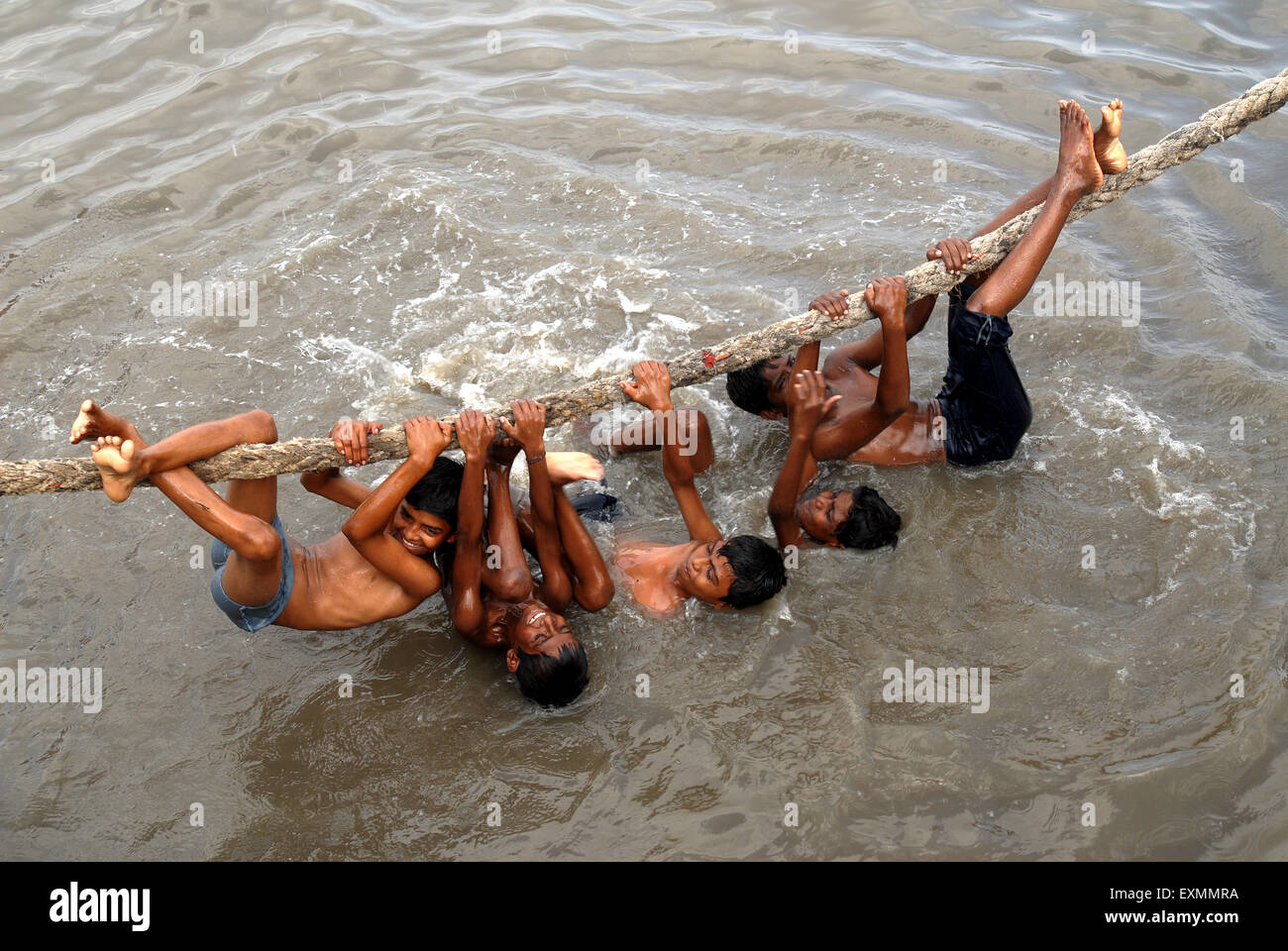 Children enjoy a dip hanging over a thick rope to beat the summer heat at Sewri creek in Bombay Mumbai ; Maharashtra Stock Photo