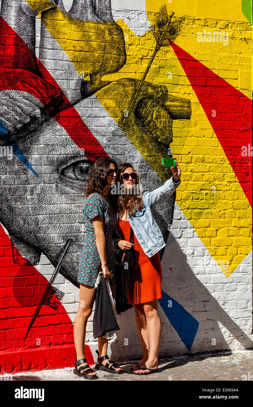 Young Women Pose For A selfie In Front Of Some Street Art/Graffiti, Brick Lane, London, England Stock Photo