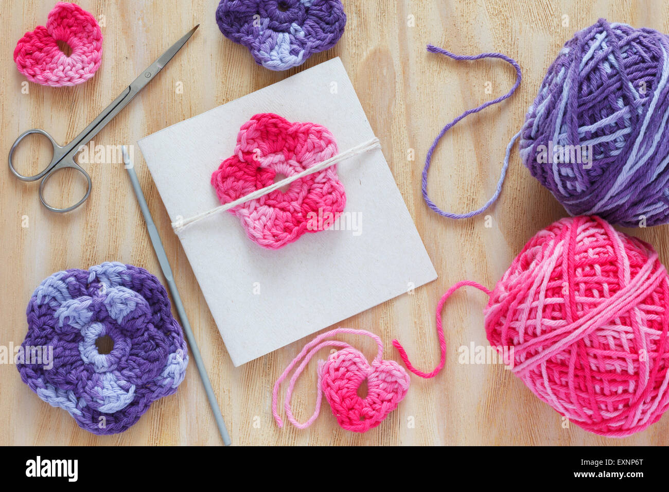 Handmade colorful crochet flowers and heart for decoration of greetings card with skein and scissors on wooden table. Selective Stock Photo