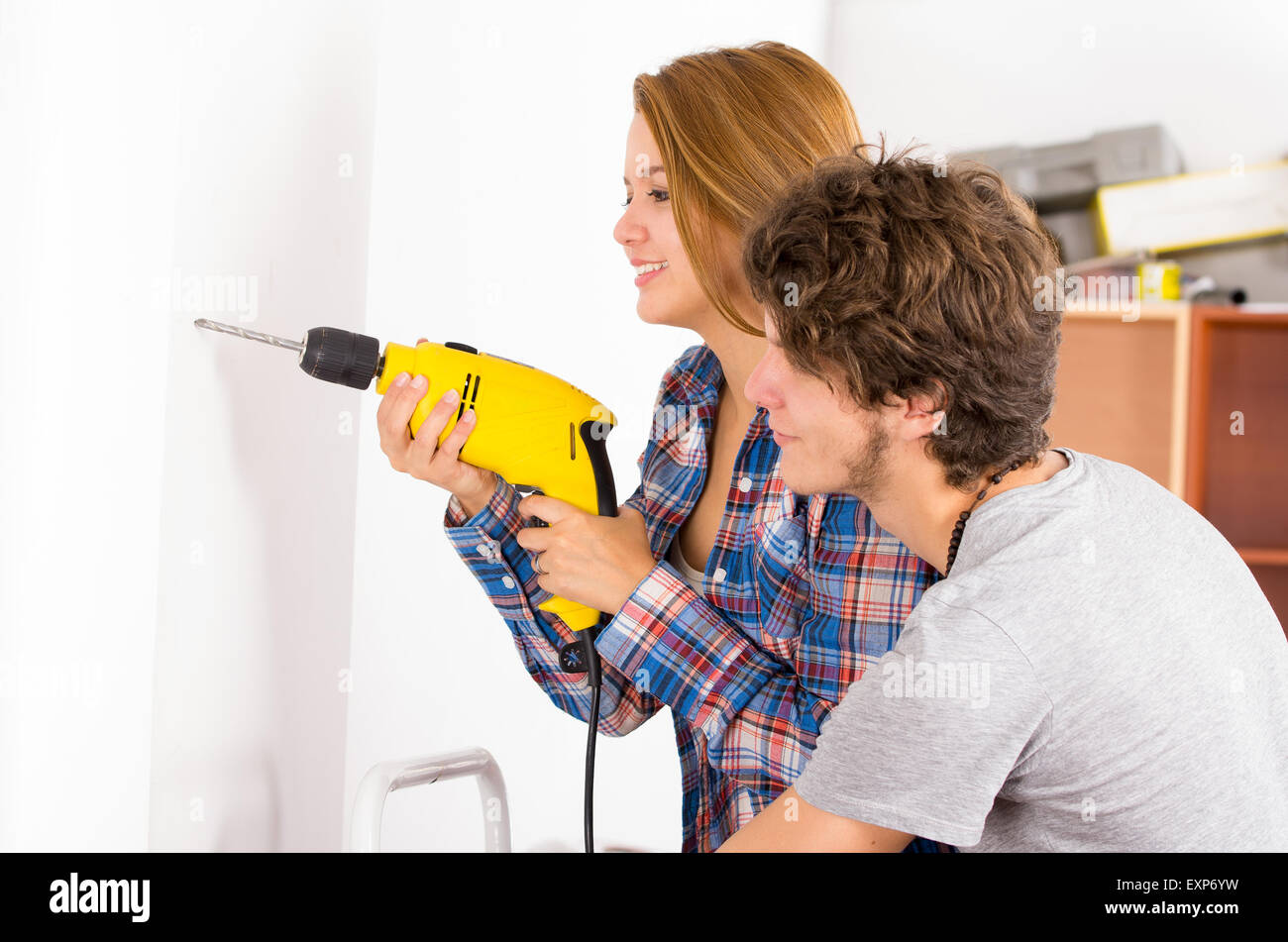 Couple renovating together as woman using power drill on wall with man standing next to her observing Stock Photo