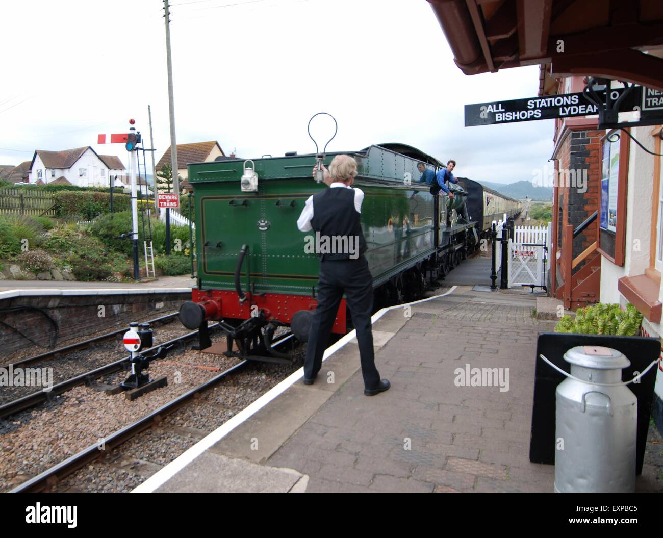 West Somerset Railway. Steam train approaching Blue Anchor station, Somerset, UK. Station guard holding the safety key aloft for collection on passing Stock Photo