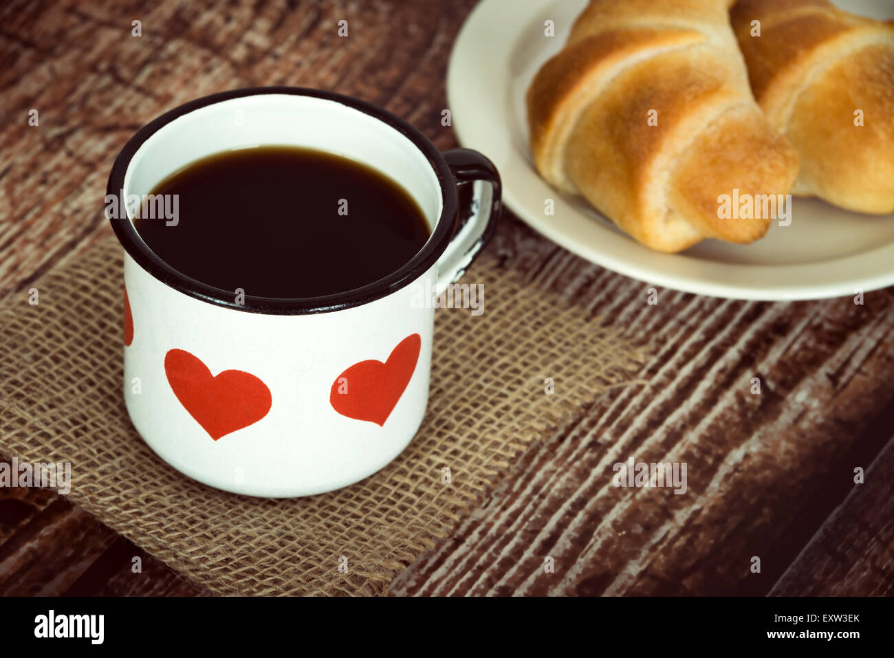 A cup of black coffee in an old enamel mug, breakfast croissants on background, vintage rustic table Stock Photo