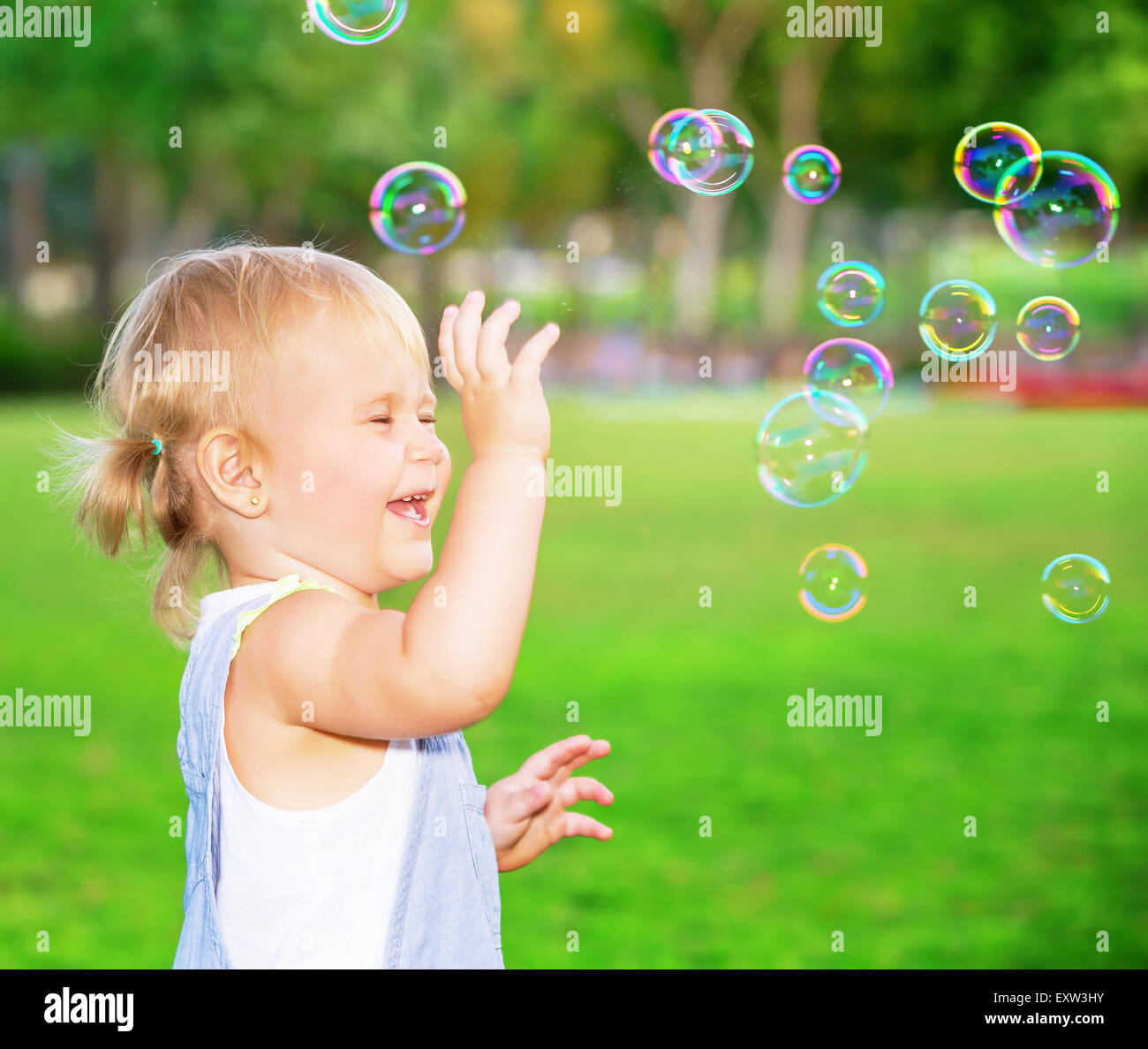 Happy child having fun in the park, cute blond baby girl playing with soap bubbles on the yard, joyful little kid Stock Photo