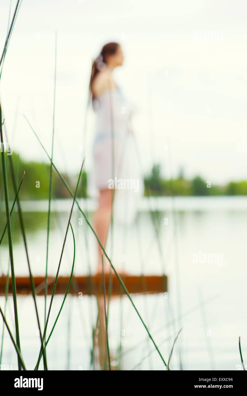Woman standing at end of lake pier enjoying view, focus on tall grass in foreground Stock Photo