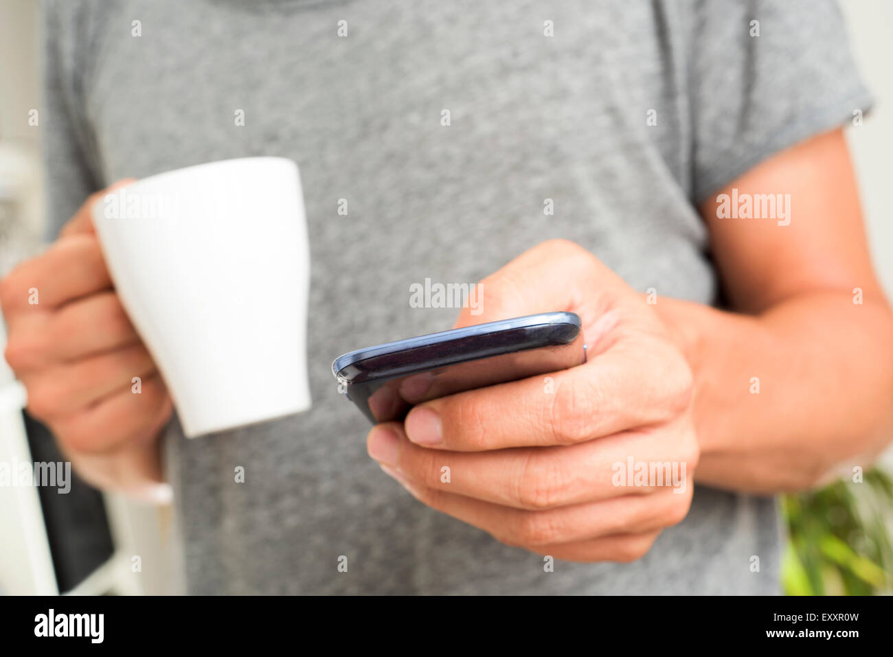 closeup of a young caucasian man in a gray tshirt using a smartphone indoors with a cup of coffee or tea in his hand Stock Photo