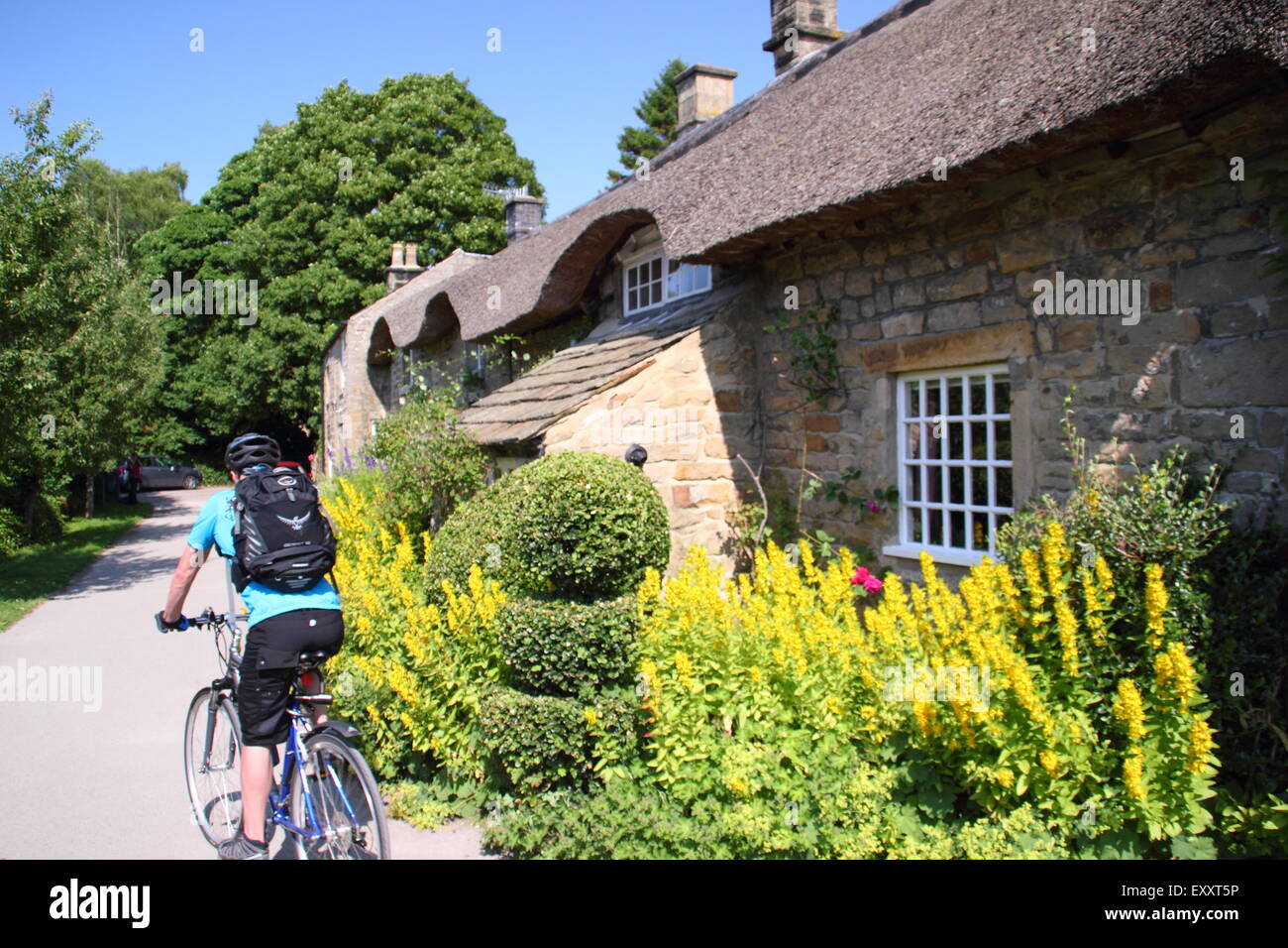 A  male cyclist  rides by a traditional English thatched cottage in Baslow village, Peak District National Park, Derbyshire, UK Stock Photo
