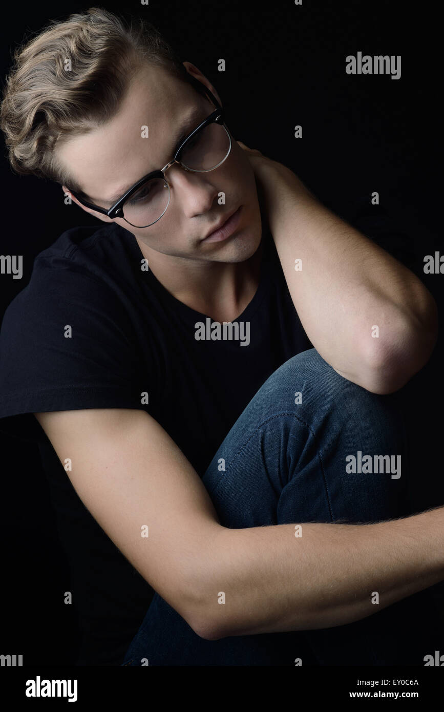 A blond wavy hair man, sitting and looking away with sadness. He appears to be lonely and emotional. A dark portrait. Stock Photo