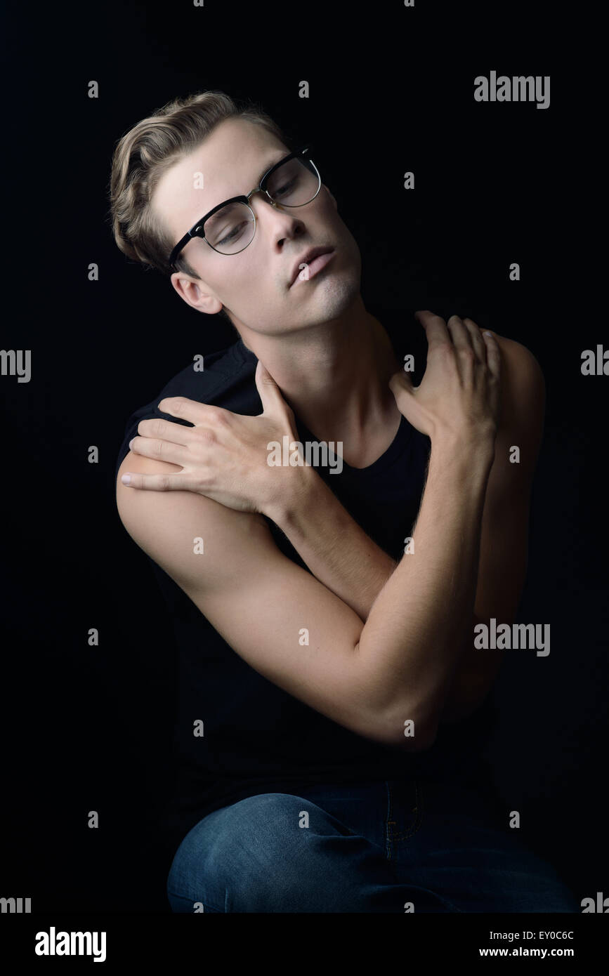 A blond man wearing vintage eye glasses, sitting and looking away with arms across his chest looking relaxed yet shy & emotional Stock Photo