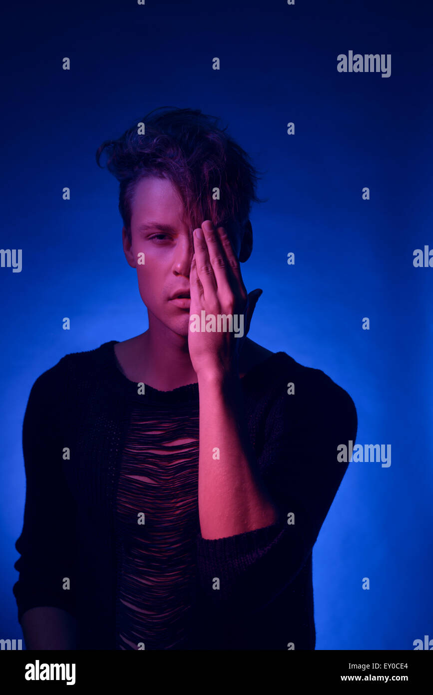 A young Caucasian male model posing with one hand covering his face, a moody high fashion dimly lit portrait concept in pink and blue light. Stock Photo