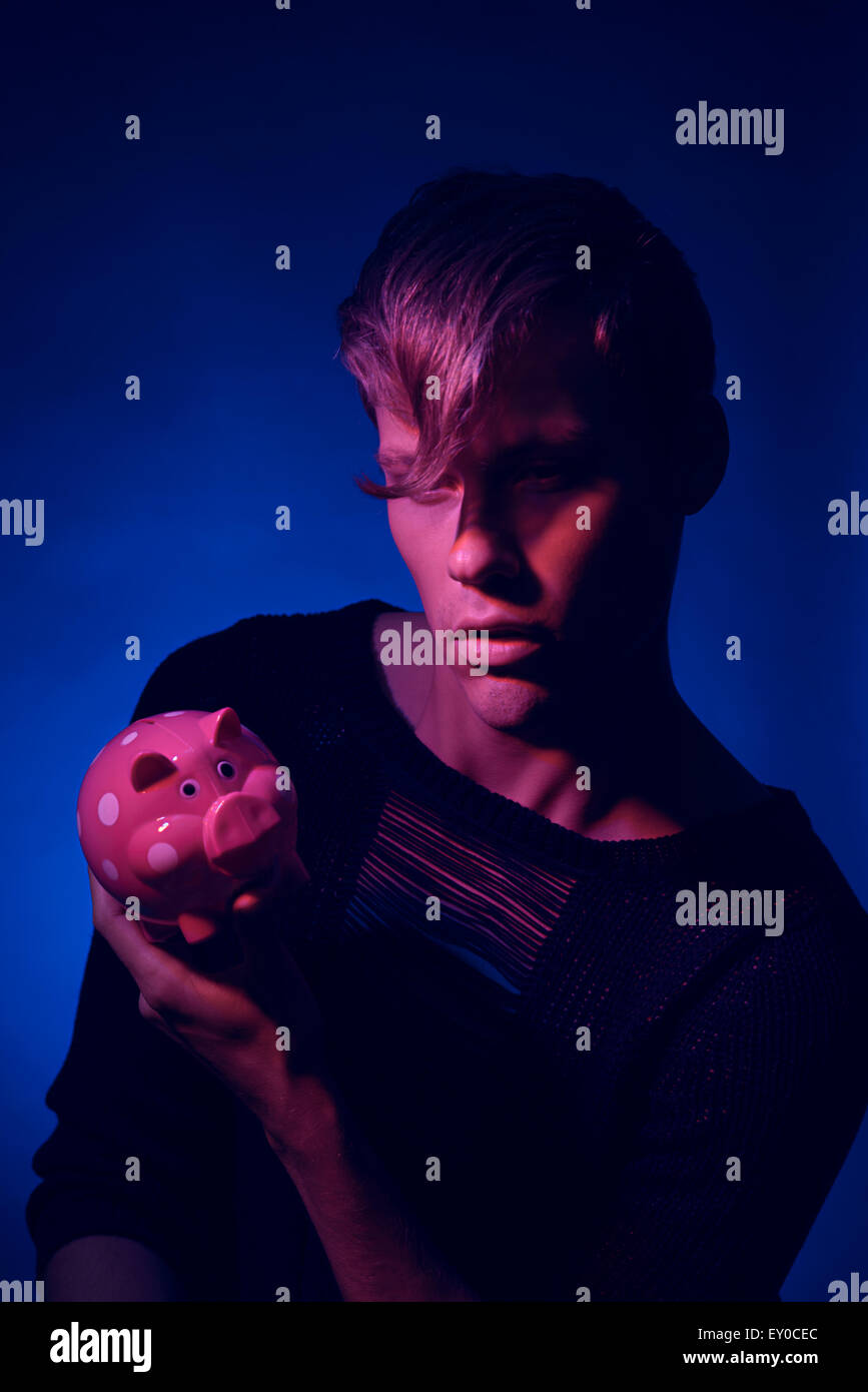 a man holding a piggy bank in his hand, a dimly lit image in pink light and blue background, a quirky fashion portrait concept. Stock Photo