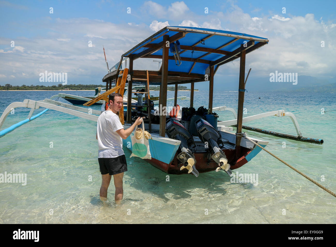 Young adullt men wearing white shirt and black shorts carrying a bag with clothes standing by the dive boat parked by the shore Stock Photo