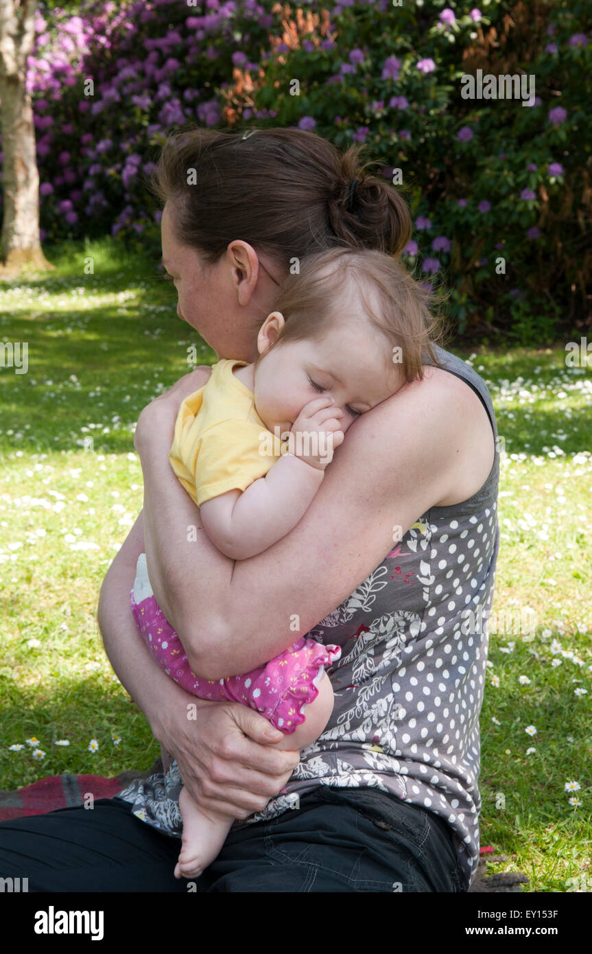 Baby girl sucking her thumb and being comforted by her mother in the the garden Stock Photo