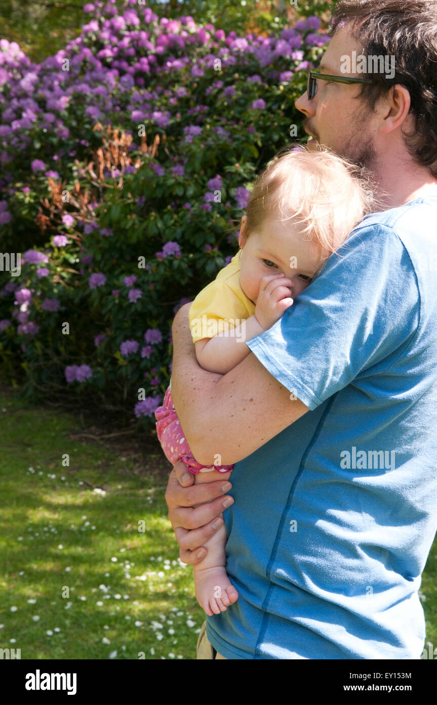 Baby girl sucking her thumb and being comforted by her father in the garden Stock Photo