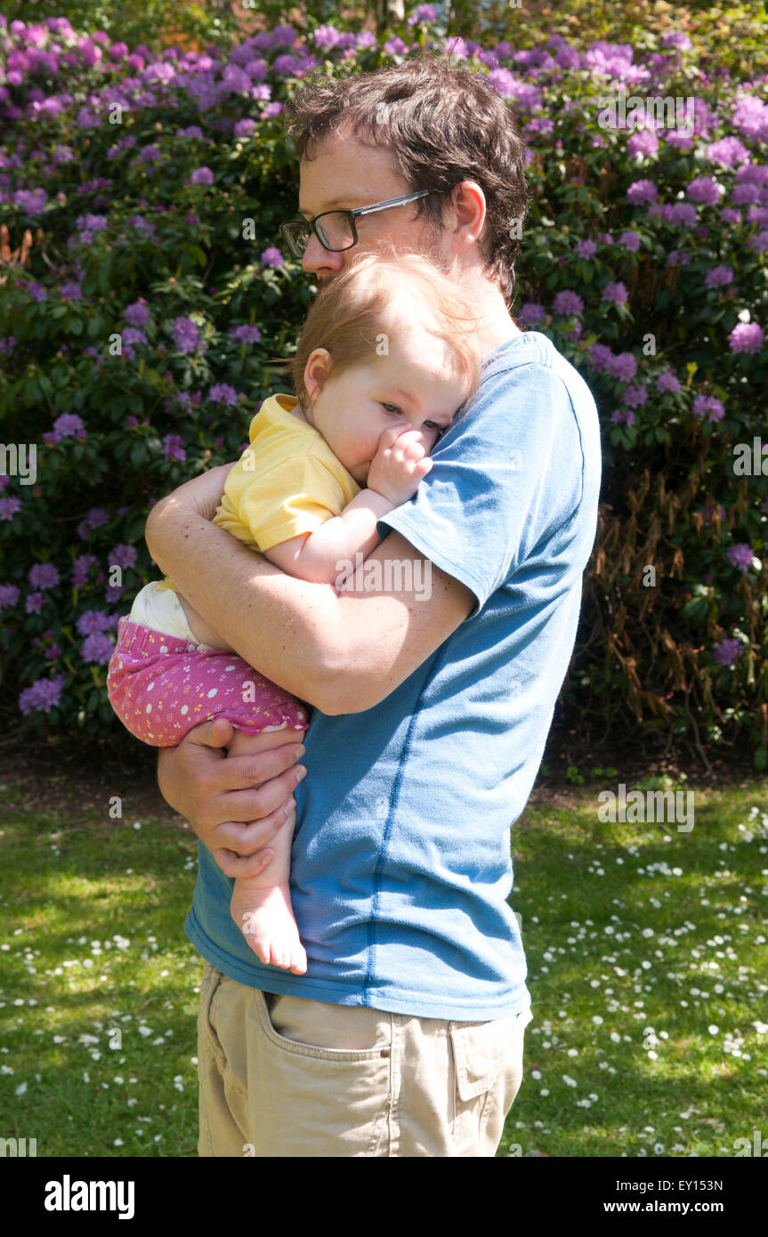 Baby girl sucking her thumb and being comforted by her father in the garden Stock Photo