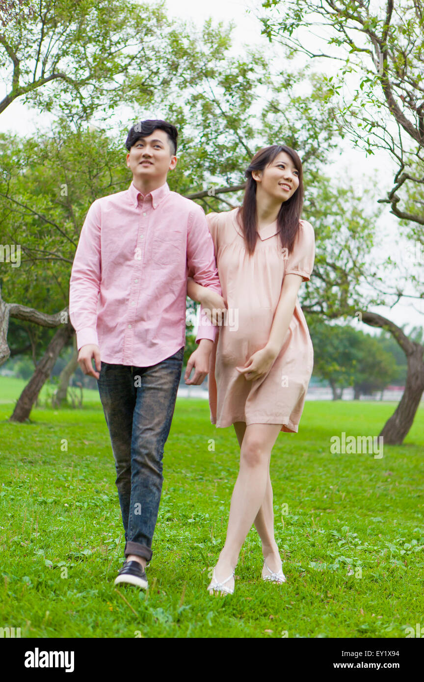 Young man and pregnant woman walking on the lawn and looking away with smile, Stock Photo