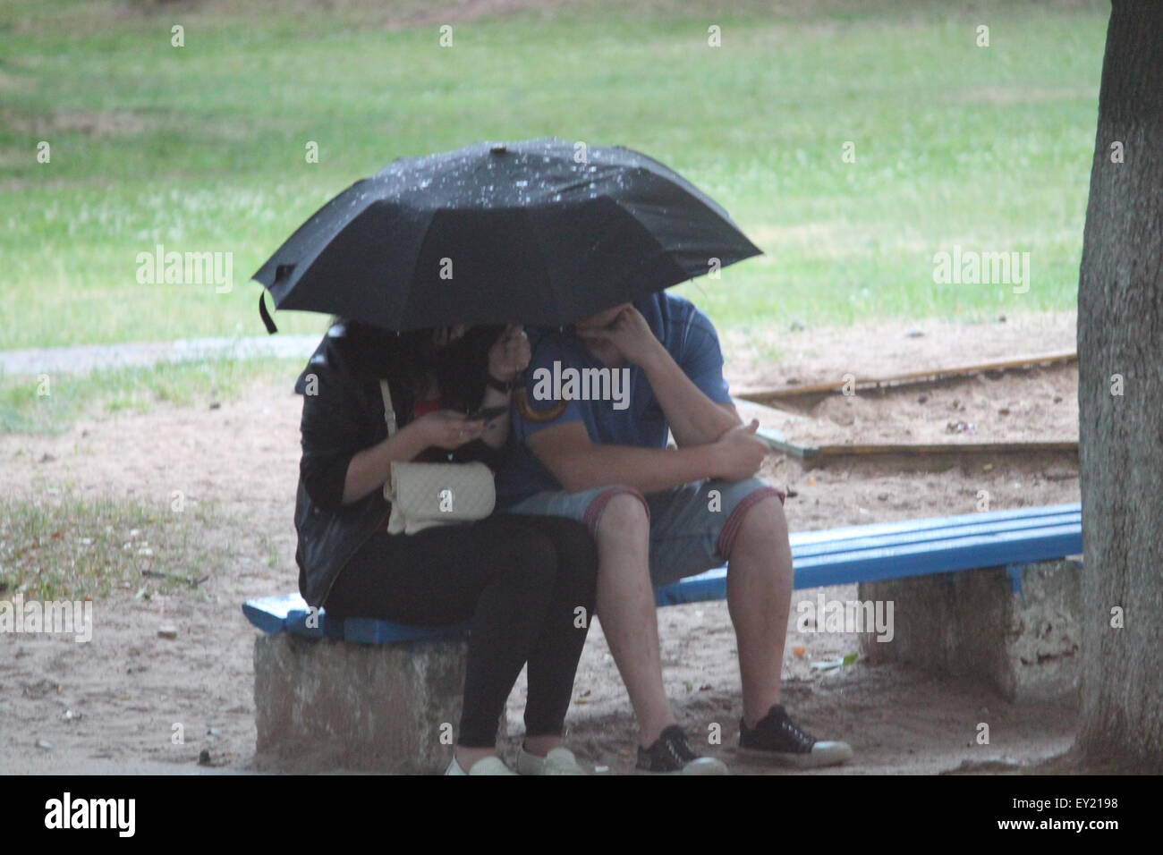 young two people sit under black umbrella under summer rain Stock Photo