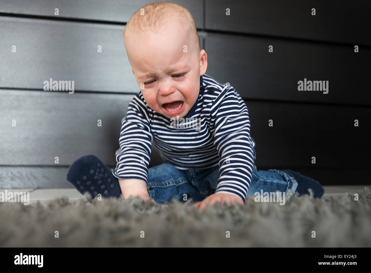 Crying baby boy sitting on rug in living room Stock Photo