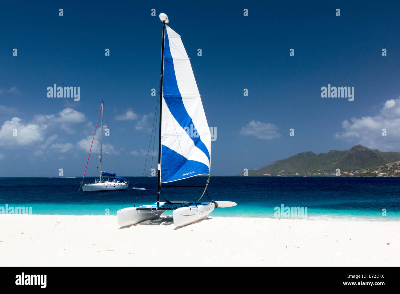 Hobie Cat with a Blue Sail on Coral Sand with Turquoise Caribbean Ocean, Moored Yacht and Union Island. Stock Photo