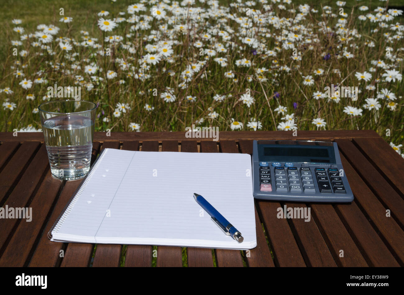 Outdoors work place with pen, paper, glass of water and calculator at a table in a garden with flowers Stock Photo