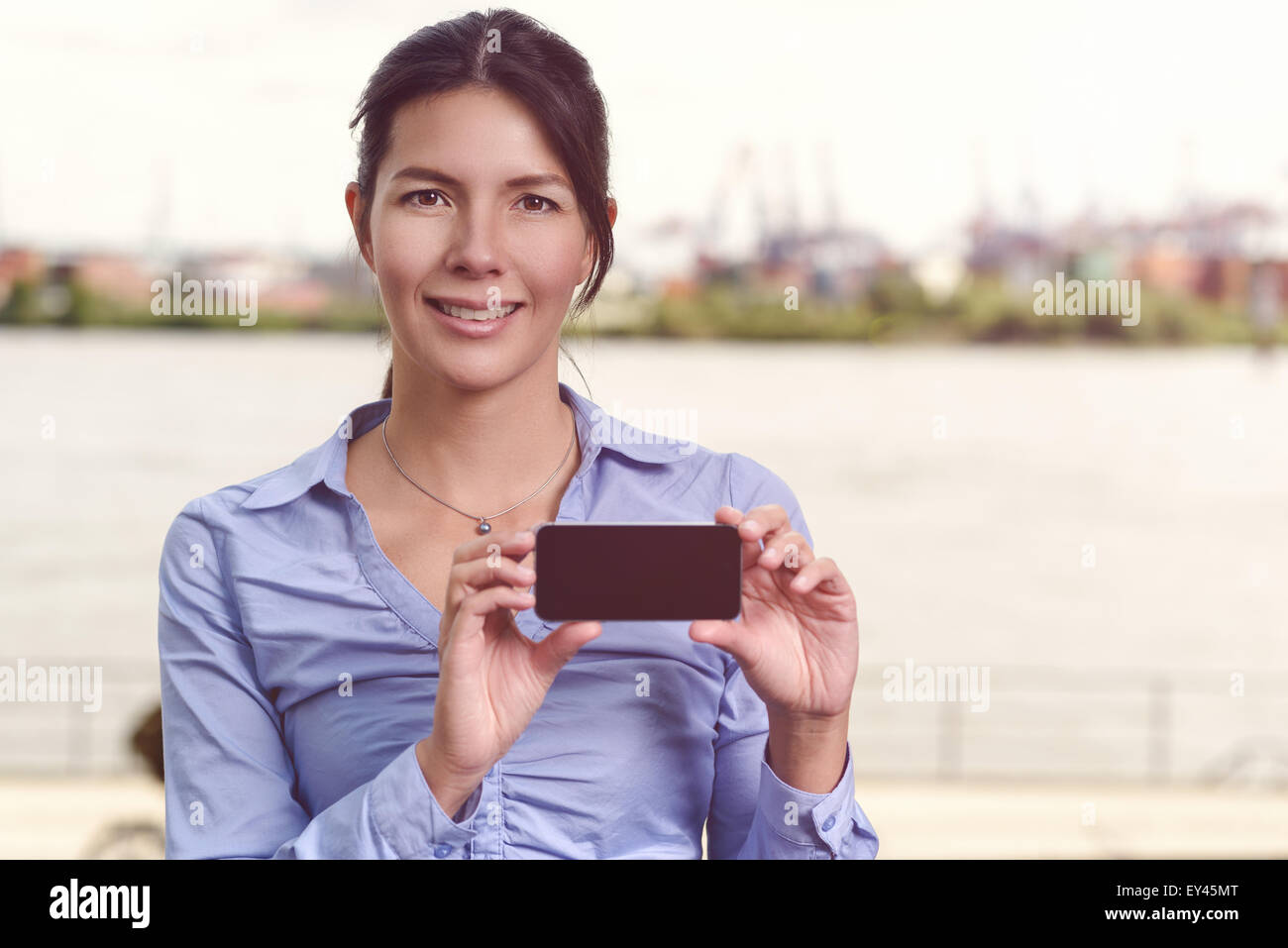 Smiling woman displaying her mobile phone for the camera with the blank screen in the horizontal position, river or waterway bac Stock Photo