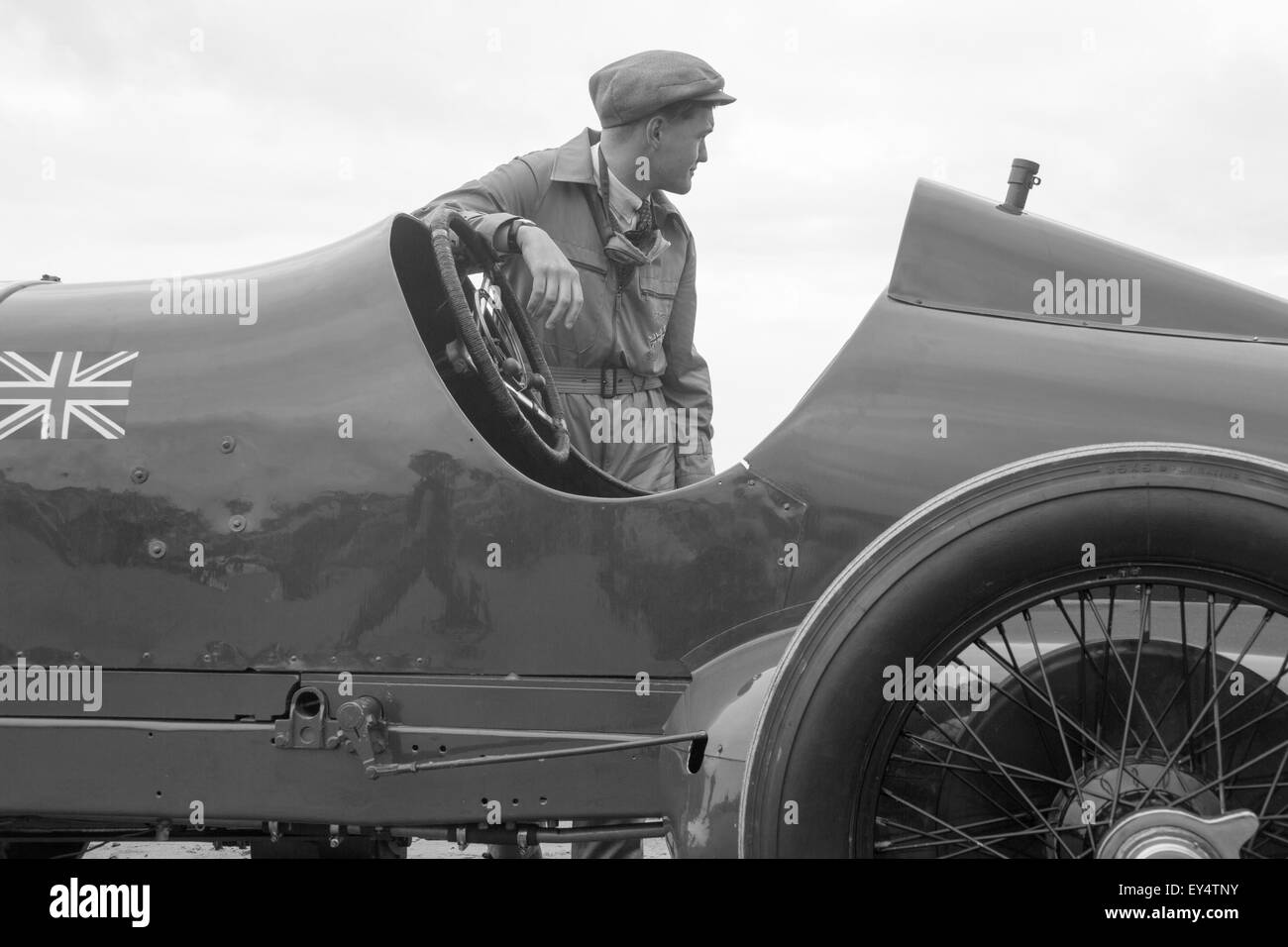 Actor dressed up in period costume with flat cap next to Bluebird racing car  steering wheel. Pictured in Black and white, on Pendine Beach, Carmarthen, Wales, UK. Stock Photo