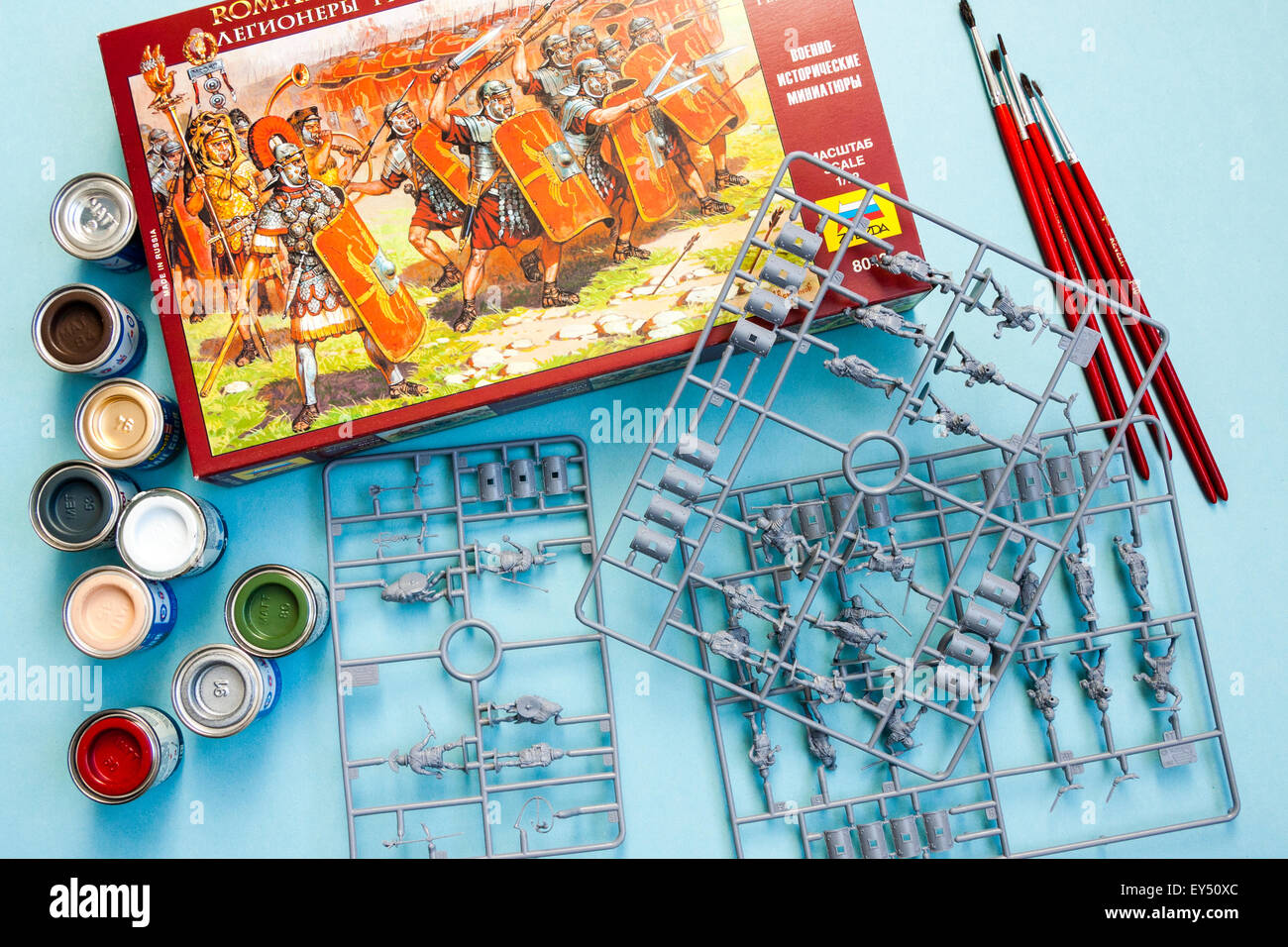 Opened box of Zvezda Ancient Roman legionaries in HO/OO plastic model figures on spurs with small paint tins and brushes on plain blue background. Stock Photo