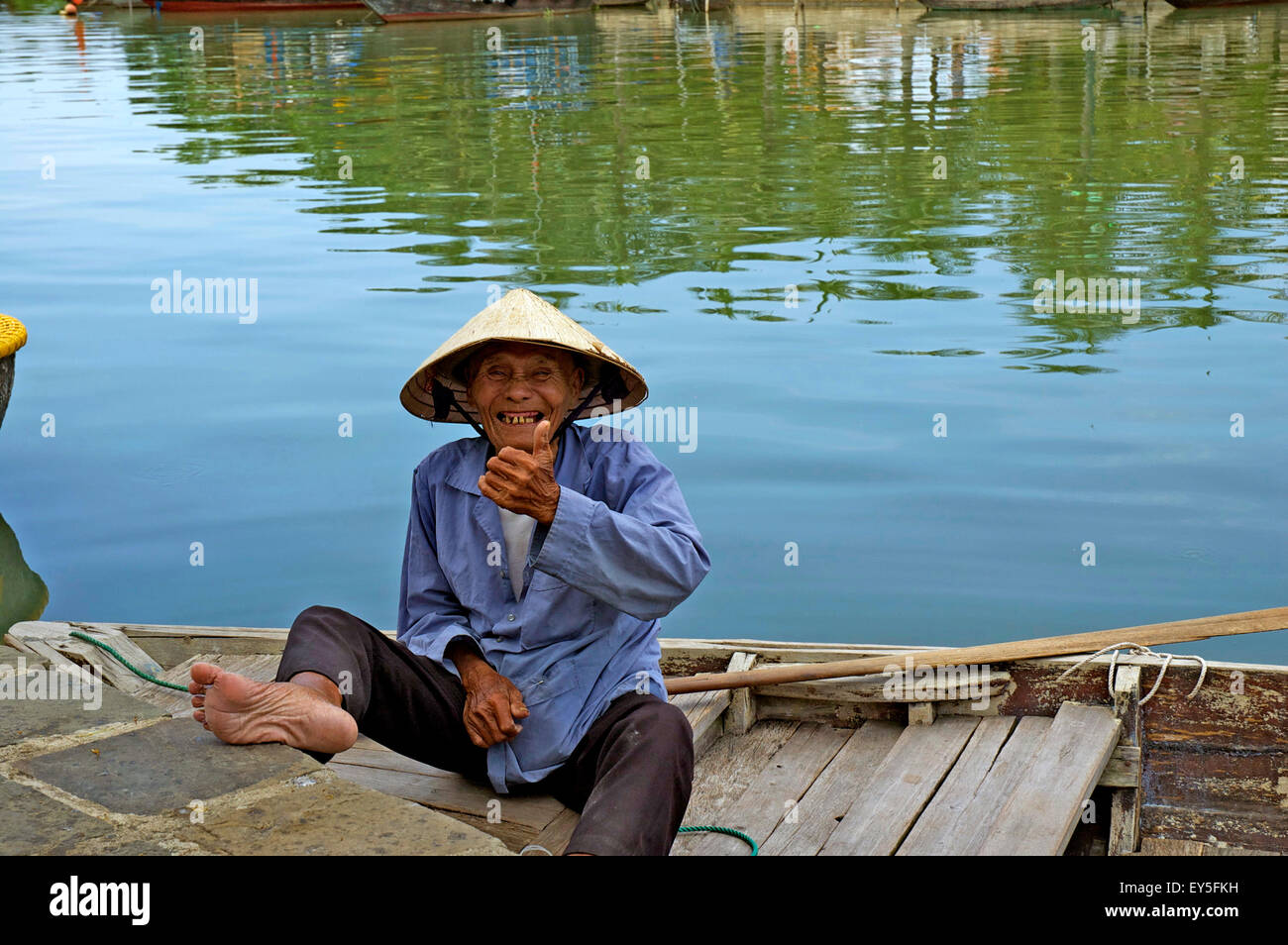An elderly Vietnamese man, wearing a traditional conical hat, smiling and giving a thumbs up sign from a boat in Hoi An. Stock Photo