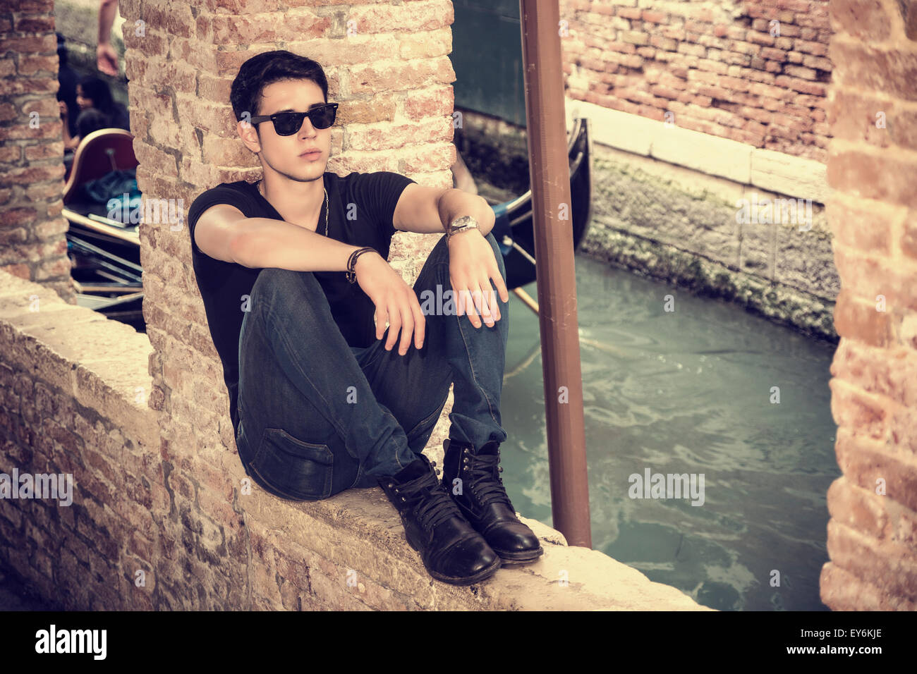 Attractive young man sitting against brick wall, looking in front of himself, wearing sunglasses Stock Photo
