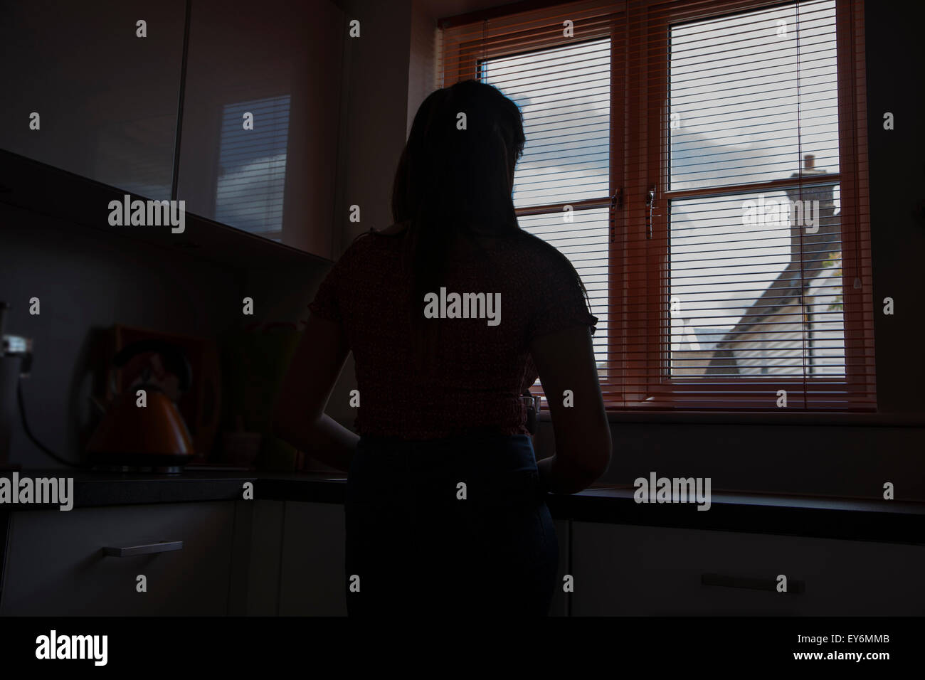 Silhouette of a young woman looking through a window blind, rear viewpoint. Stock Photo