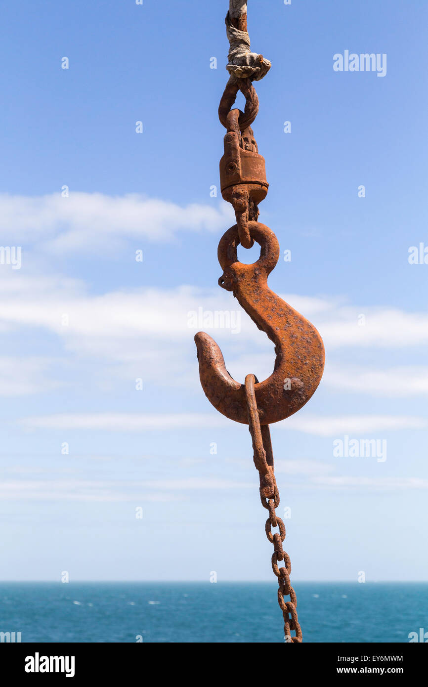 The shackled rusted hook of a crane is shown against the blue sky and sea of Portland Bill in Dorset, UK. Stock Photo