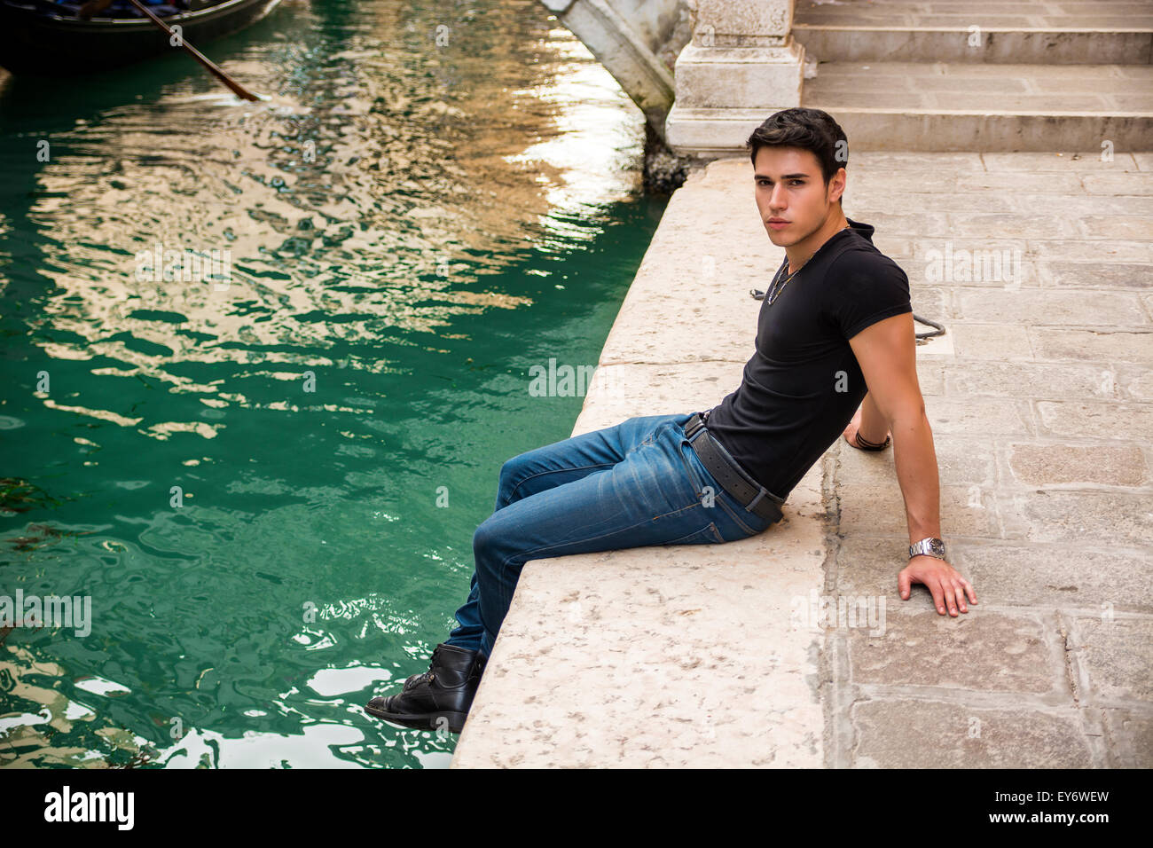 Portrait of Attractive Dark Haired Young Man Sitting Docks Next to Narrow Canal in Venice, Italy Stock Photo
