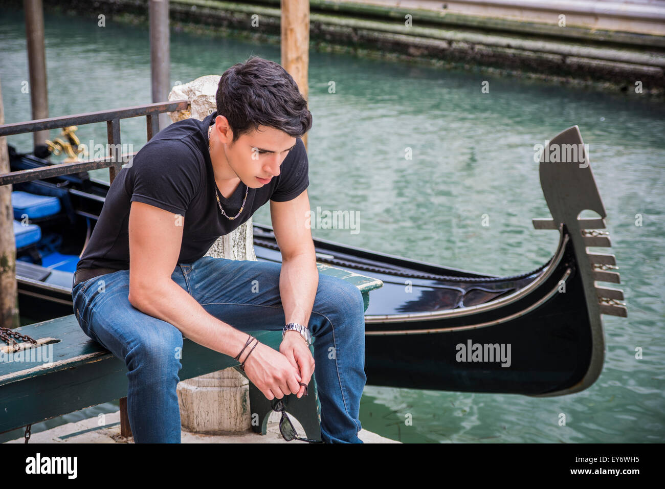 Portrait of Attractive Dark Haired Young Man Sitting on Bench Next to Narrow Canal in Venice, Italy, with Traditional Gondola Stock Photo