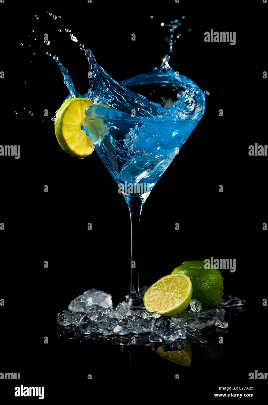 Ice cube making a splash in martini glass with blue cocktail, garnished with lime slice, lime, crushed ice. Black background. Stock Photo