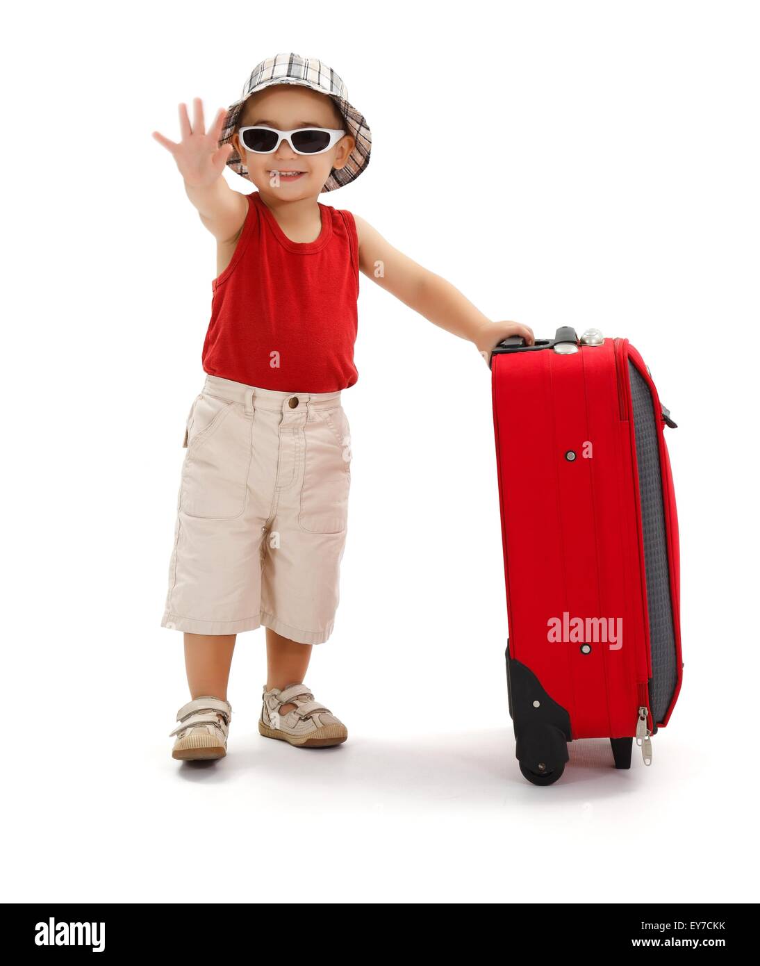 Child standing near luggage, wearing hat and sunglasses, holding his luggage and waving good bye with hand Stock Photo