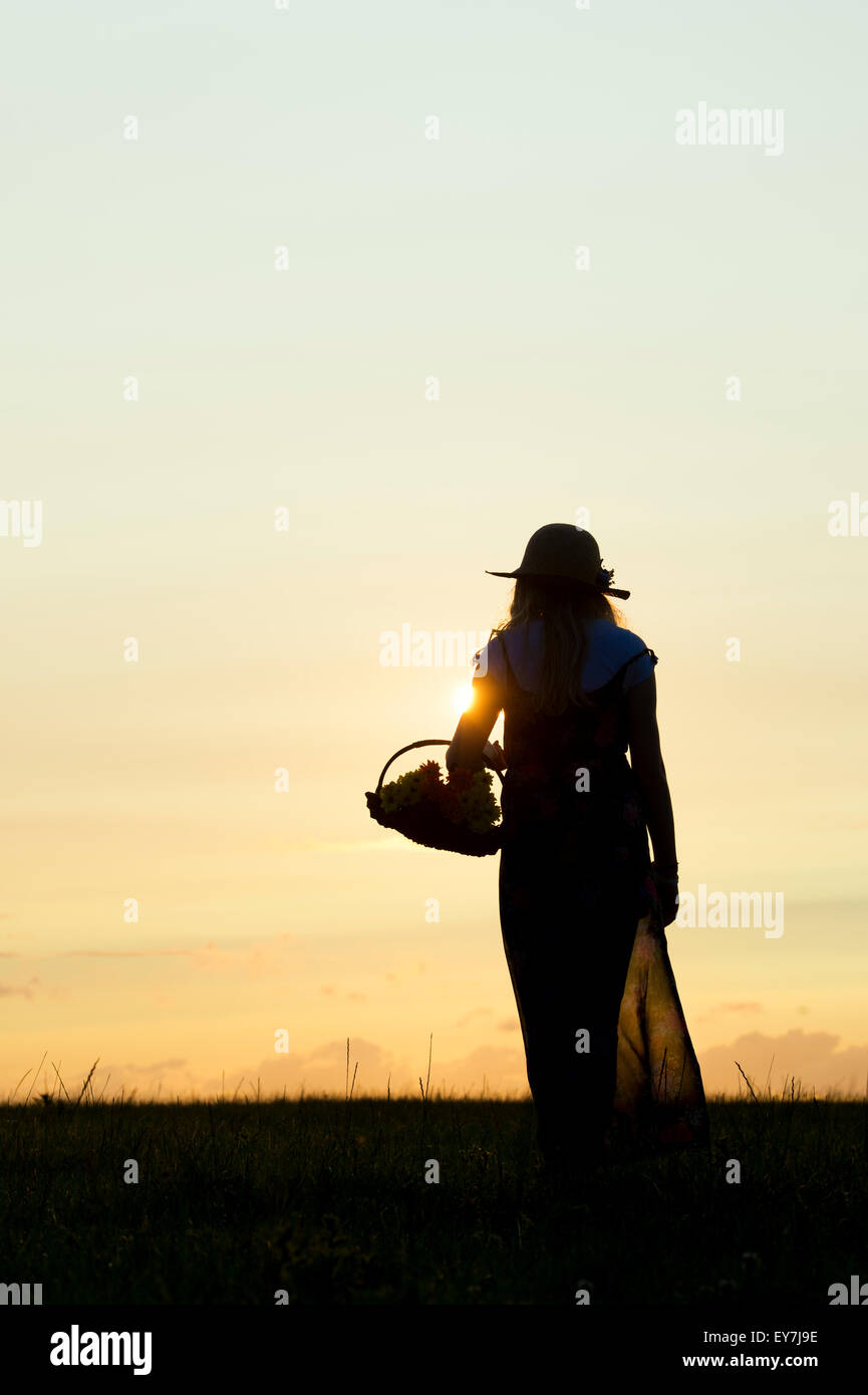 Teenage girl wearing a summer hat walking holding a basket of flowers at sunset. Silhouette Stock Photo