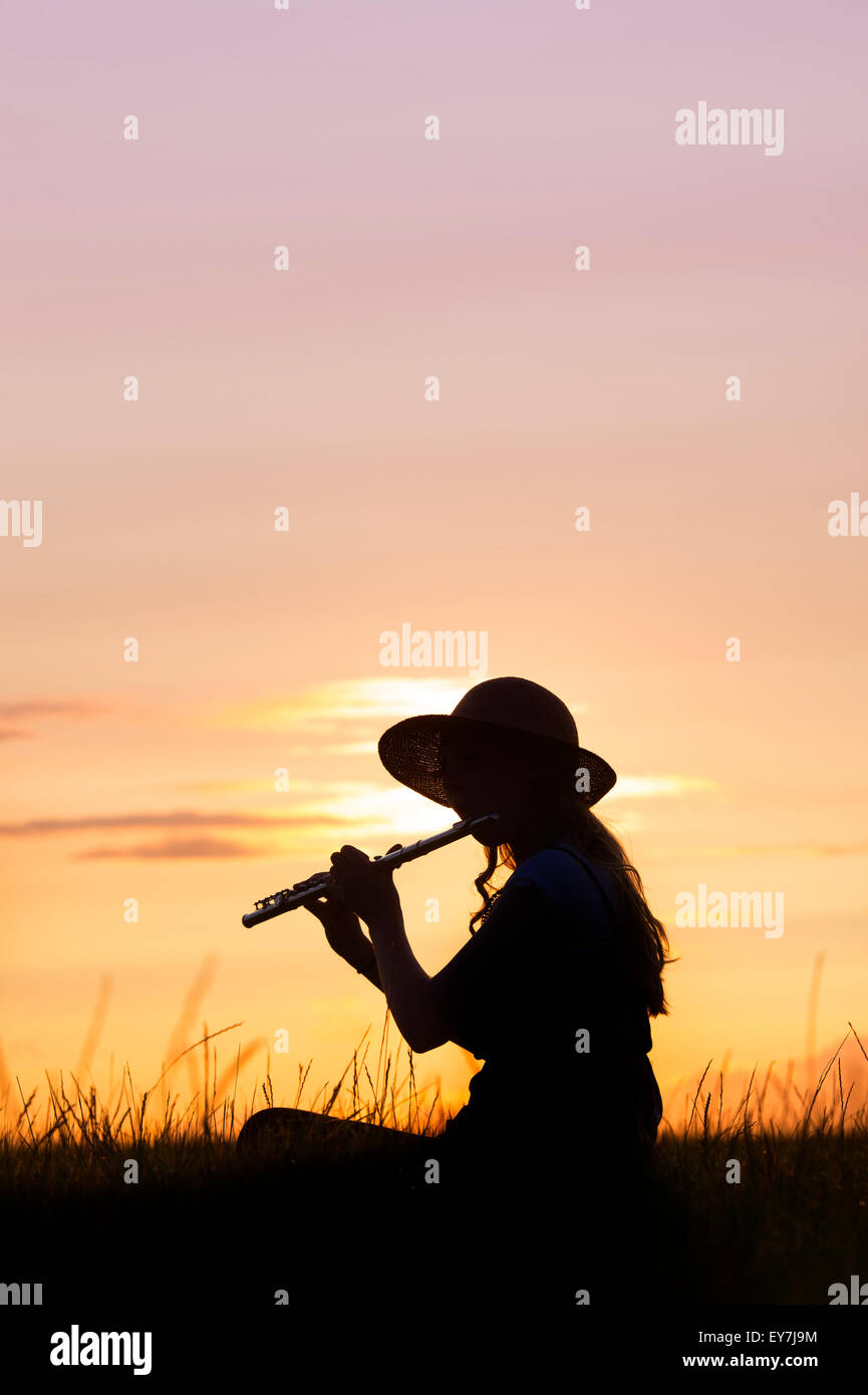 Teenage girl sat in the grass playing a flute at sunset. Silhouette Stock Photo