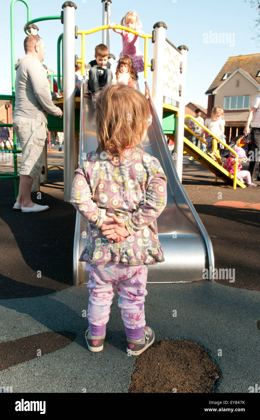 Rear view of toddler holding her hands behind her back looking at children playing on a climbing frame Stock Photo