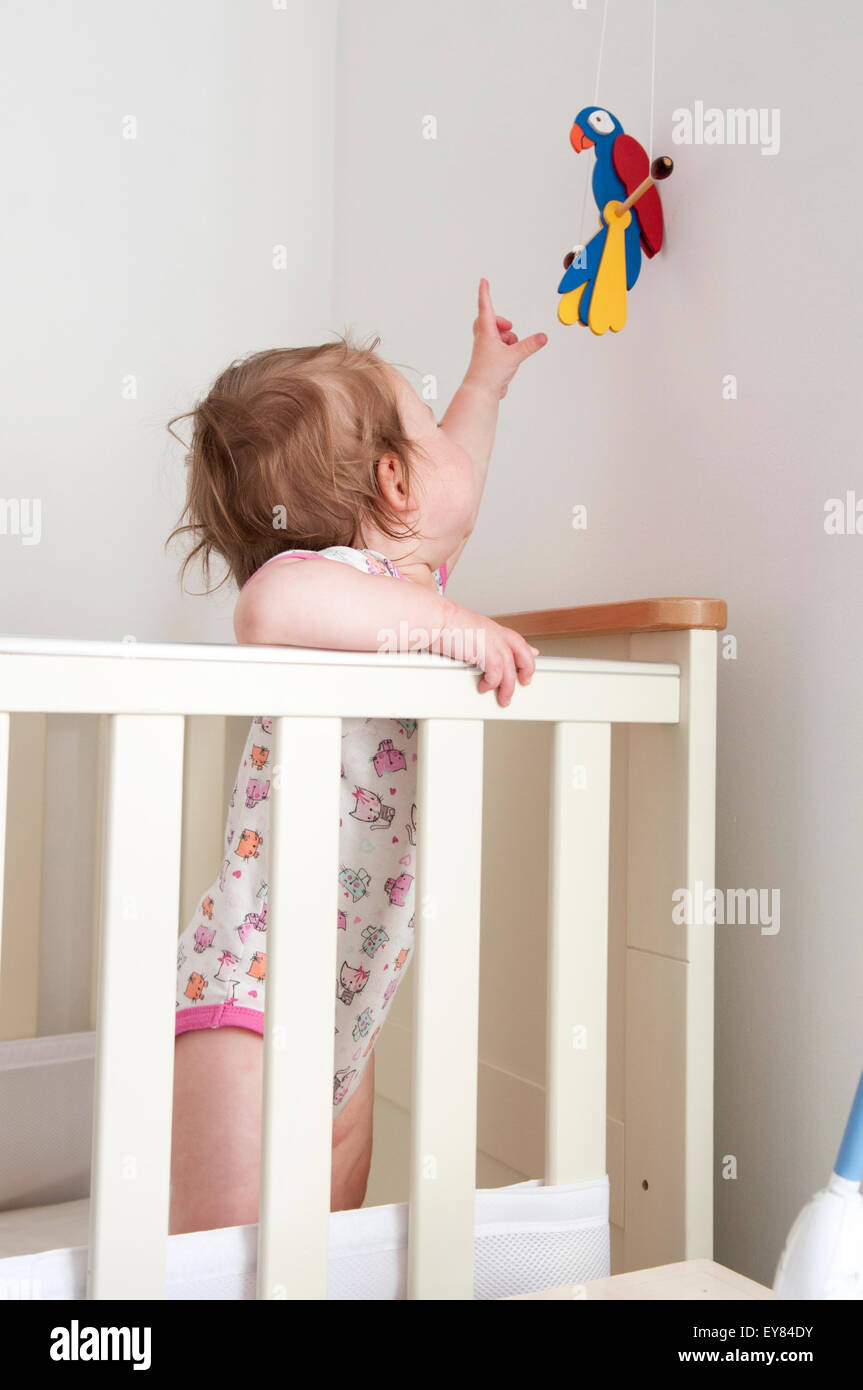 Baby girl standing inside her cot reaching for a hanging toy Stock Photo