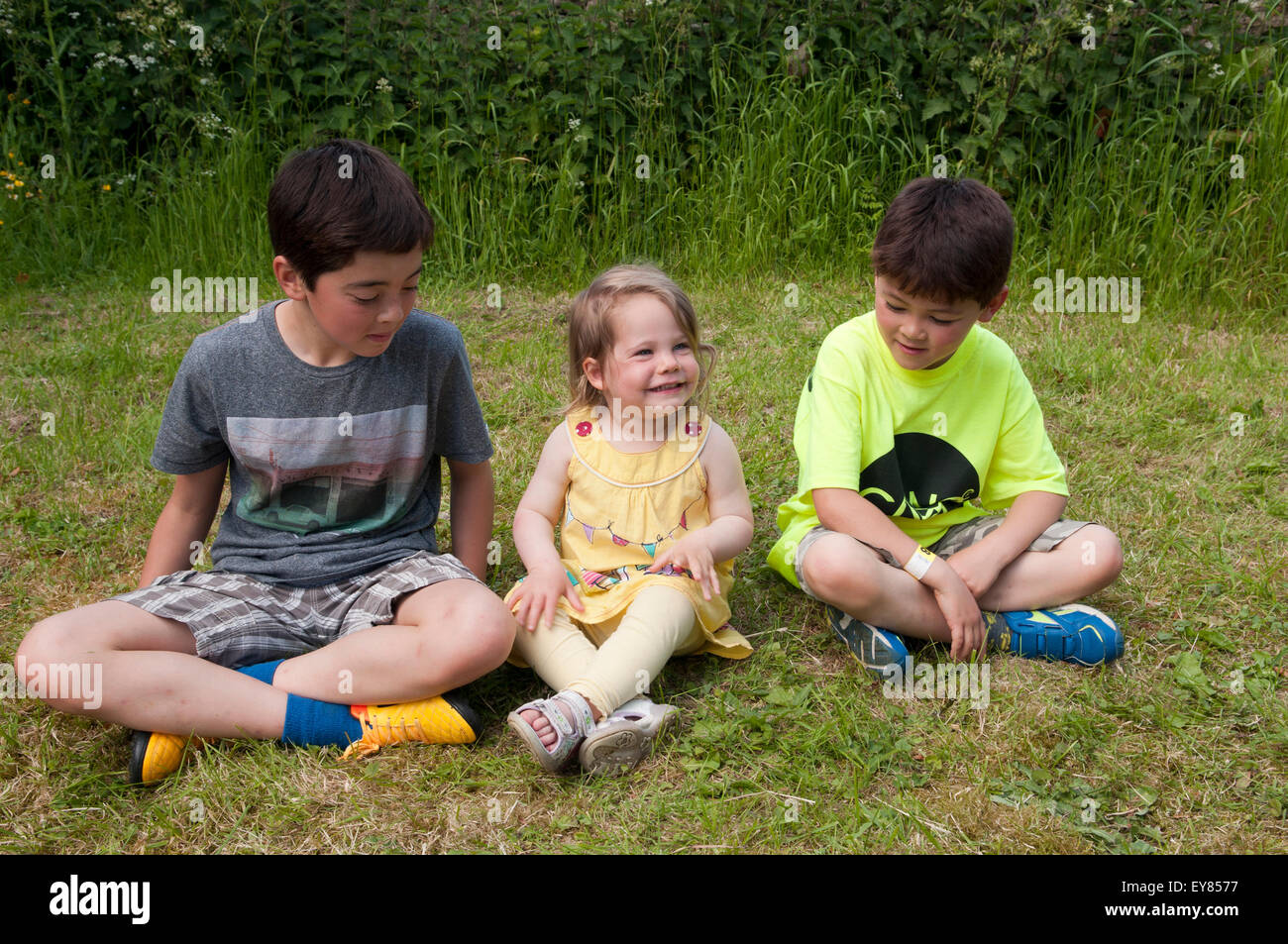 Little girl smiling, sitting on the grass with two boys Stock Photo