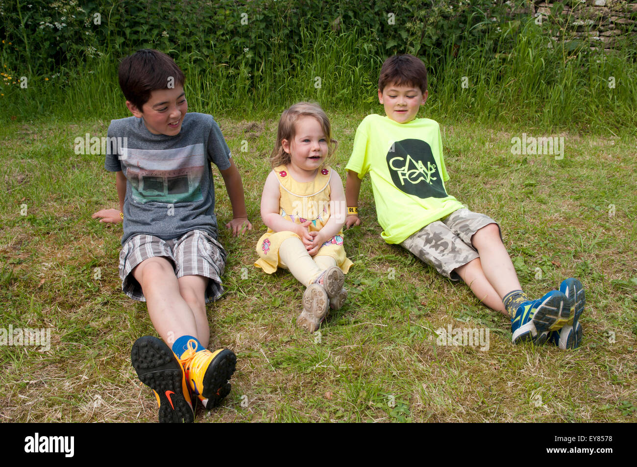 Little girl smiling, sitting on the grass with two boys Stock Photo