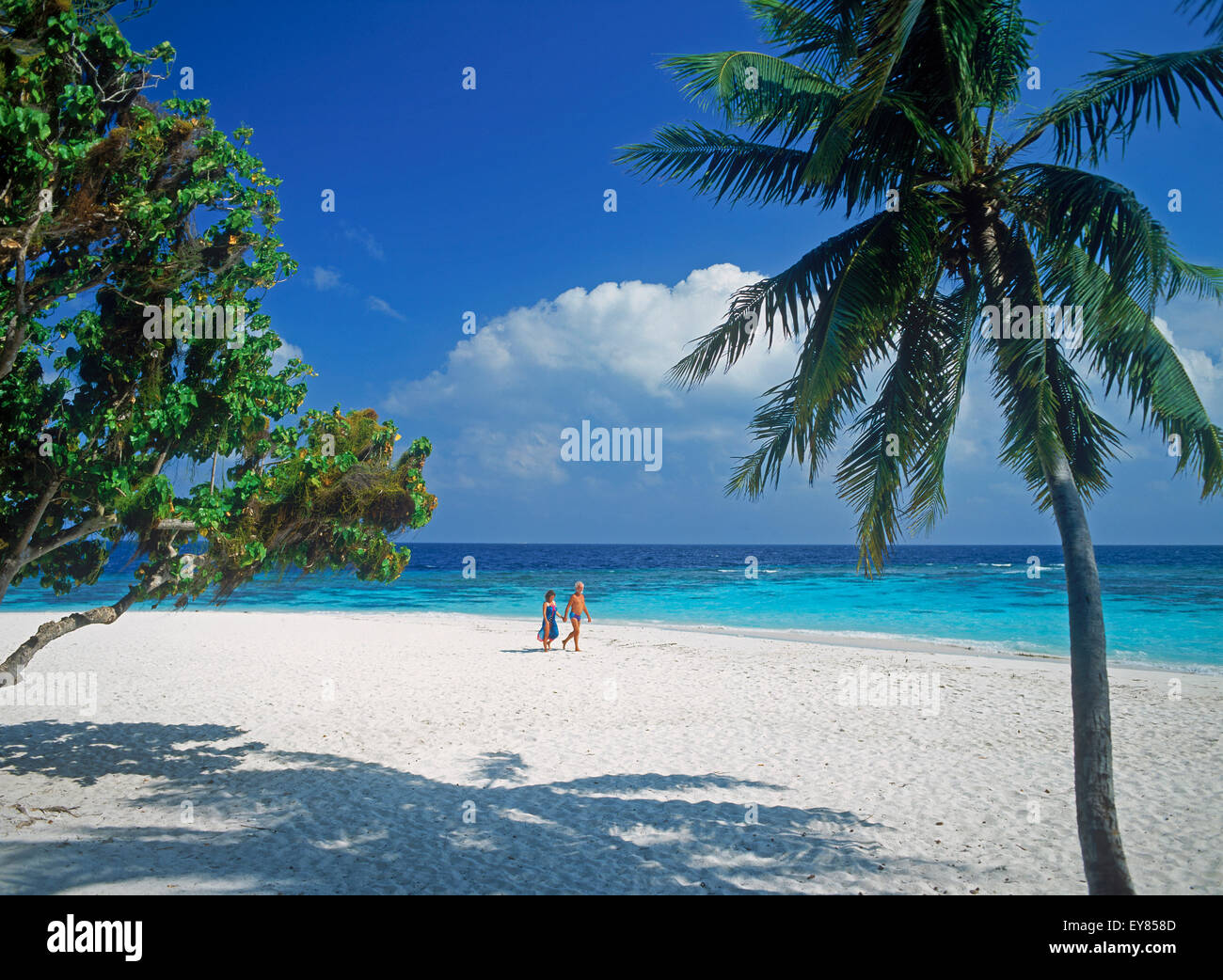 Couple walking on tropical island in Maldives across white sands under blue skies next to clear aqua waters Stock Photo